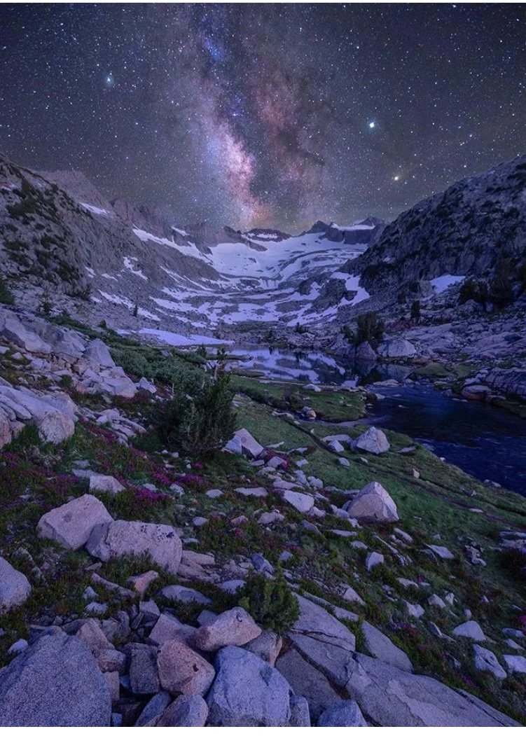 Nighttime mountain landscape with snow patches, a flowing river, and a starry sky featuring the Milky Way galaxy.