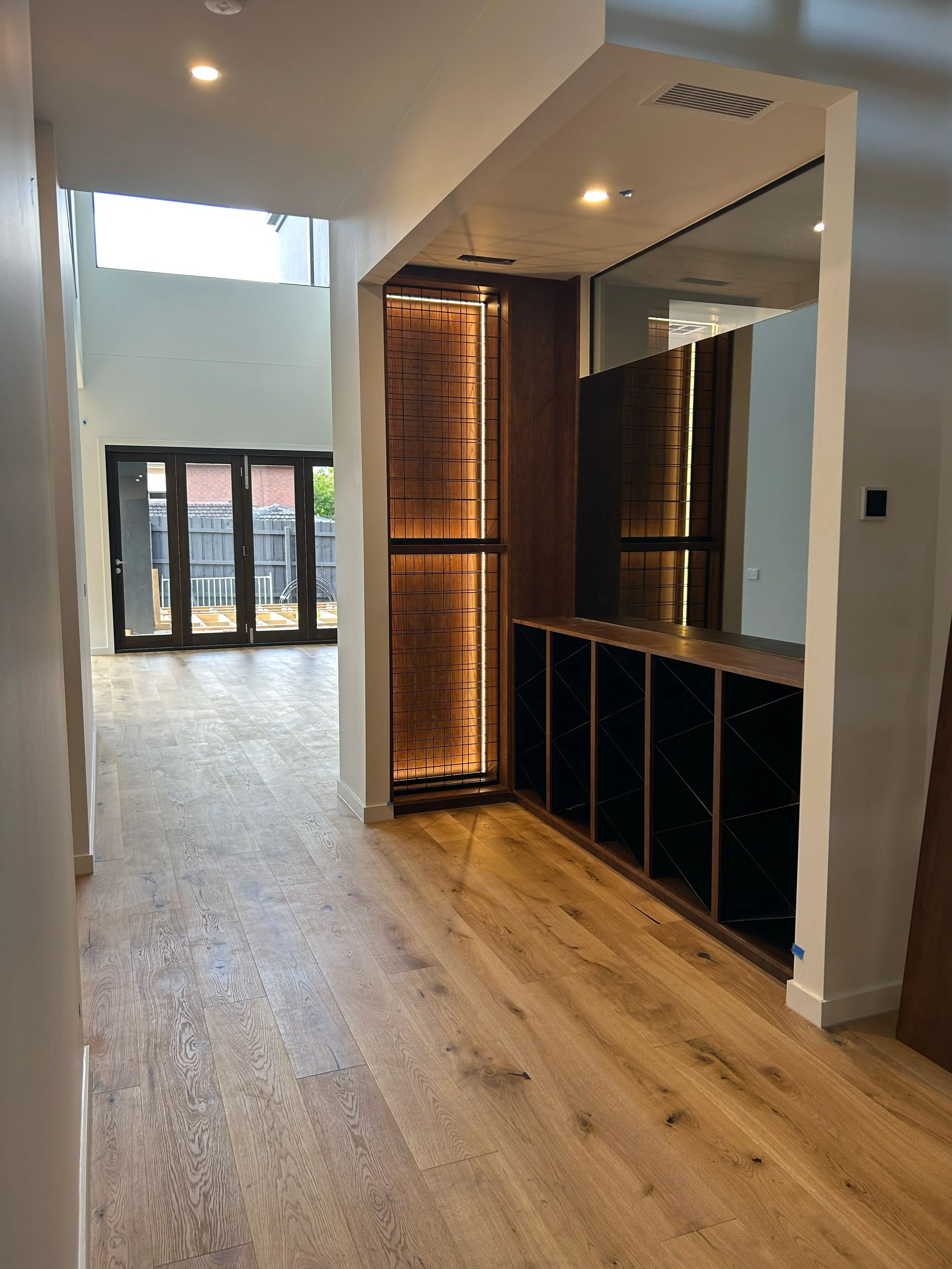 Interior view of a modern home with light wood flooring, white walls, and a built-in wooden cabinet with black storage compartments, illuminated by warm LED lighting, with a large mirror on the right side and glass doors leading outside.