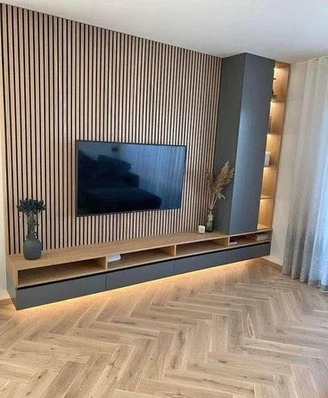 Modern living room with wooden slat accent wall, mounted flat-screen TV, gray and wood TV console, decorative vases, and built-in shelving with lighting.