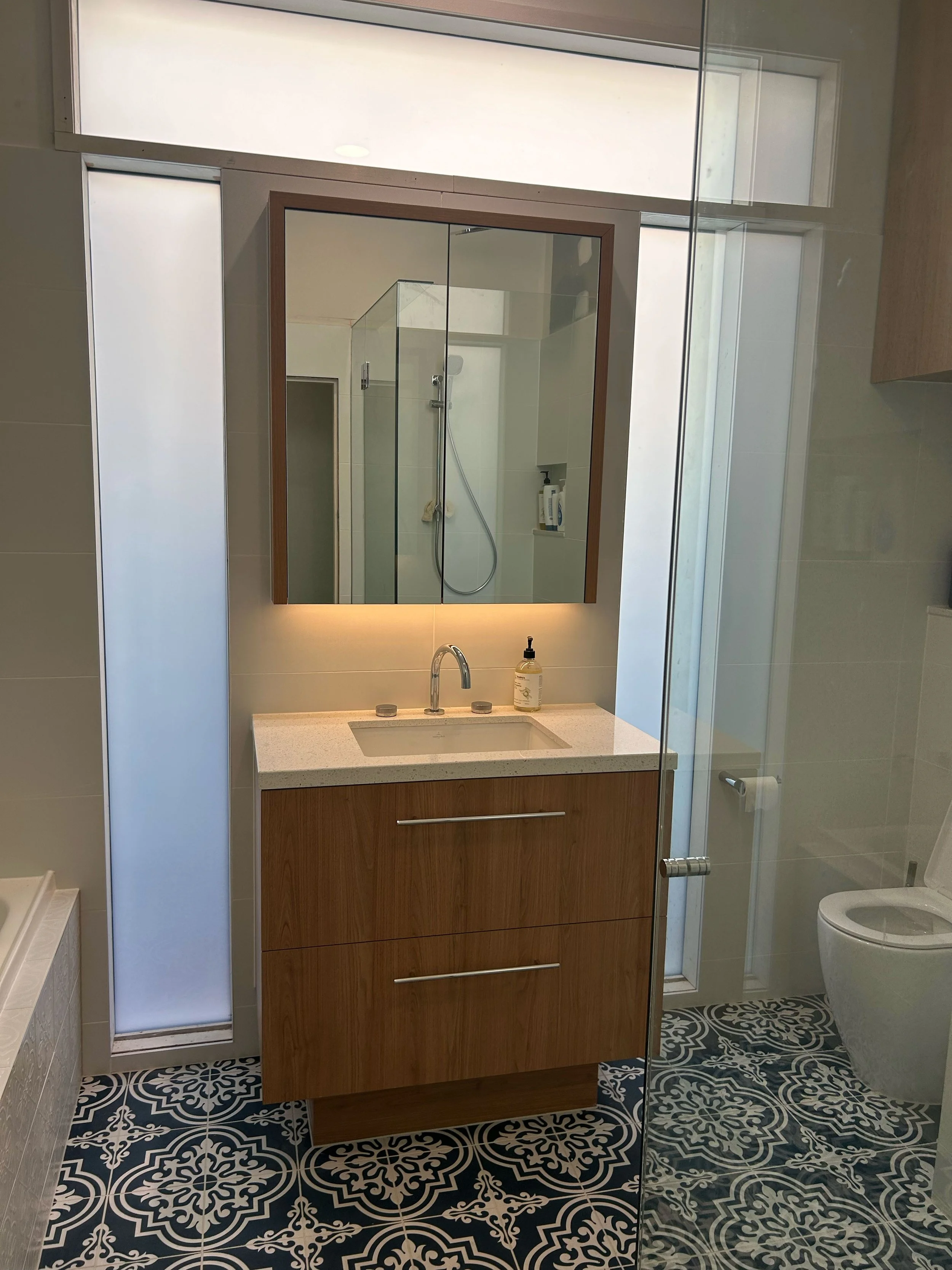 Modern bathroom with a wooden vanity, large mirror, frosted glass panels, a shower with a glass door, and a toilet with patterned black and white floor tiles.