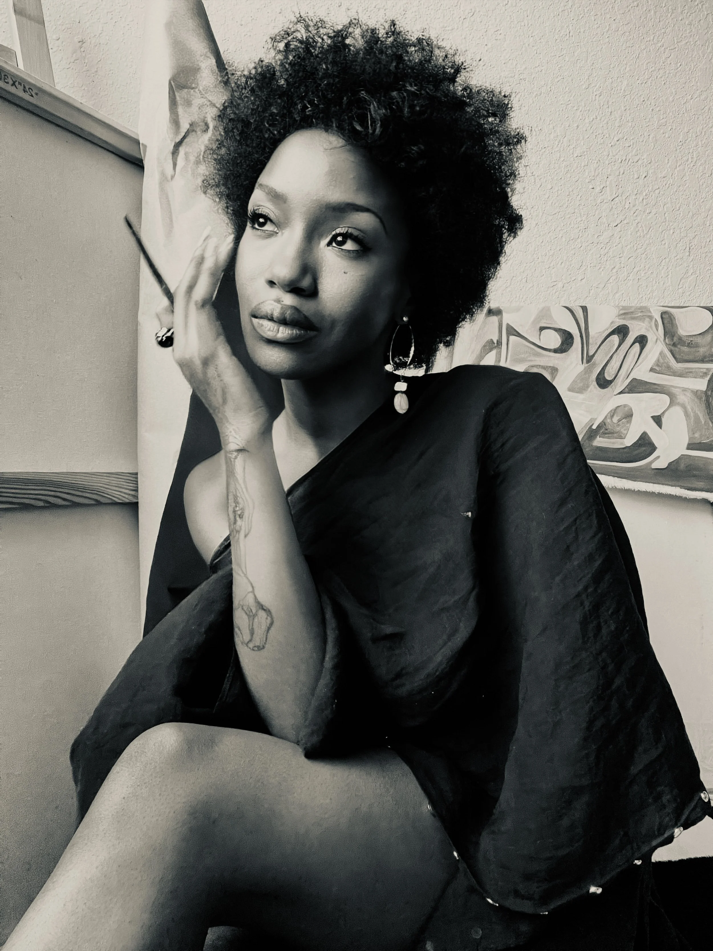 Black and white photograph of a woman with natural curly hair, sitting with her legs crossed, gazing thoughtfully. She is wearing earrings, a dark loose top, and has an arm tattoo. She is holding a cigarette near her face, with background artwork visible.