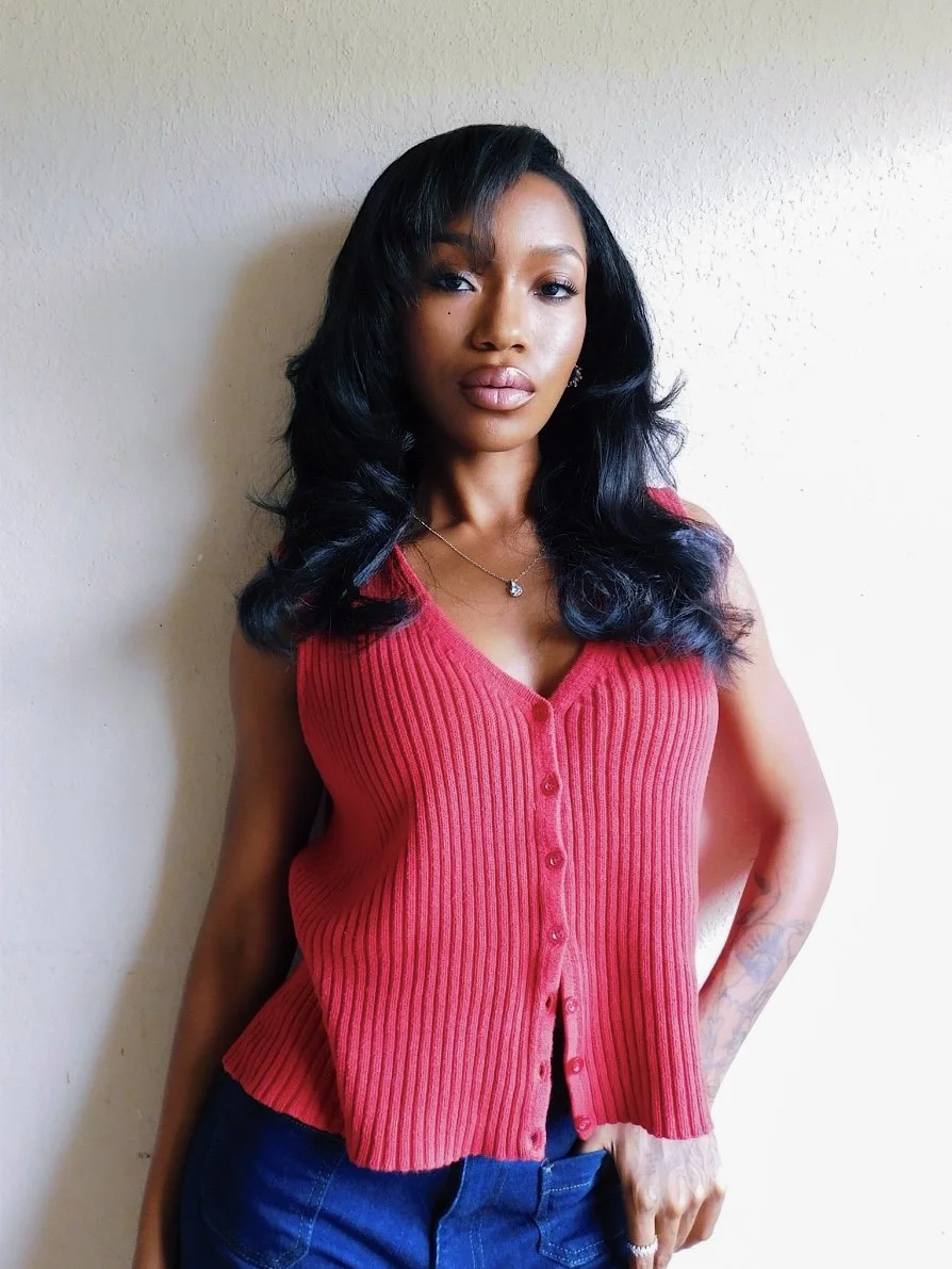 A woman with long dark hair wearing a red sleeveless ribbed button-up top and dark pants stands against a plain light-colored wall.
