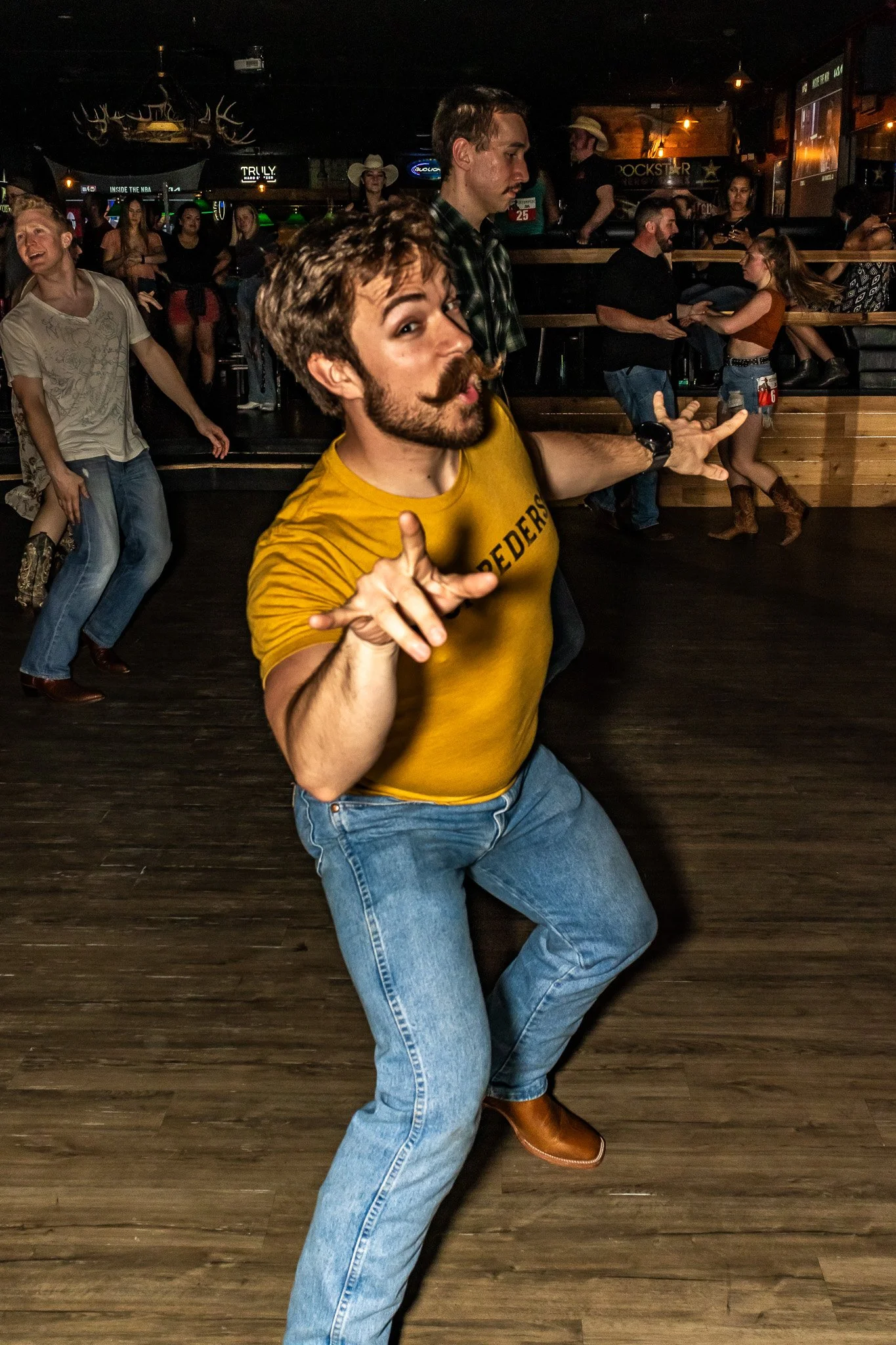 Man dancing in a bar wearing a yellow shirt and blue jeans, with people in the background also dancing and socializing.