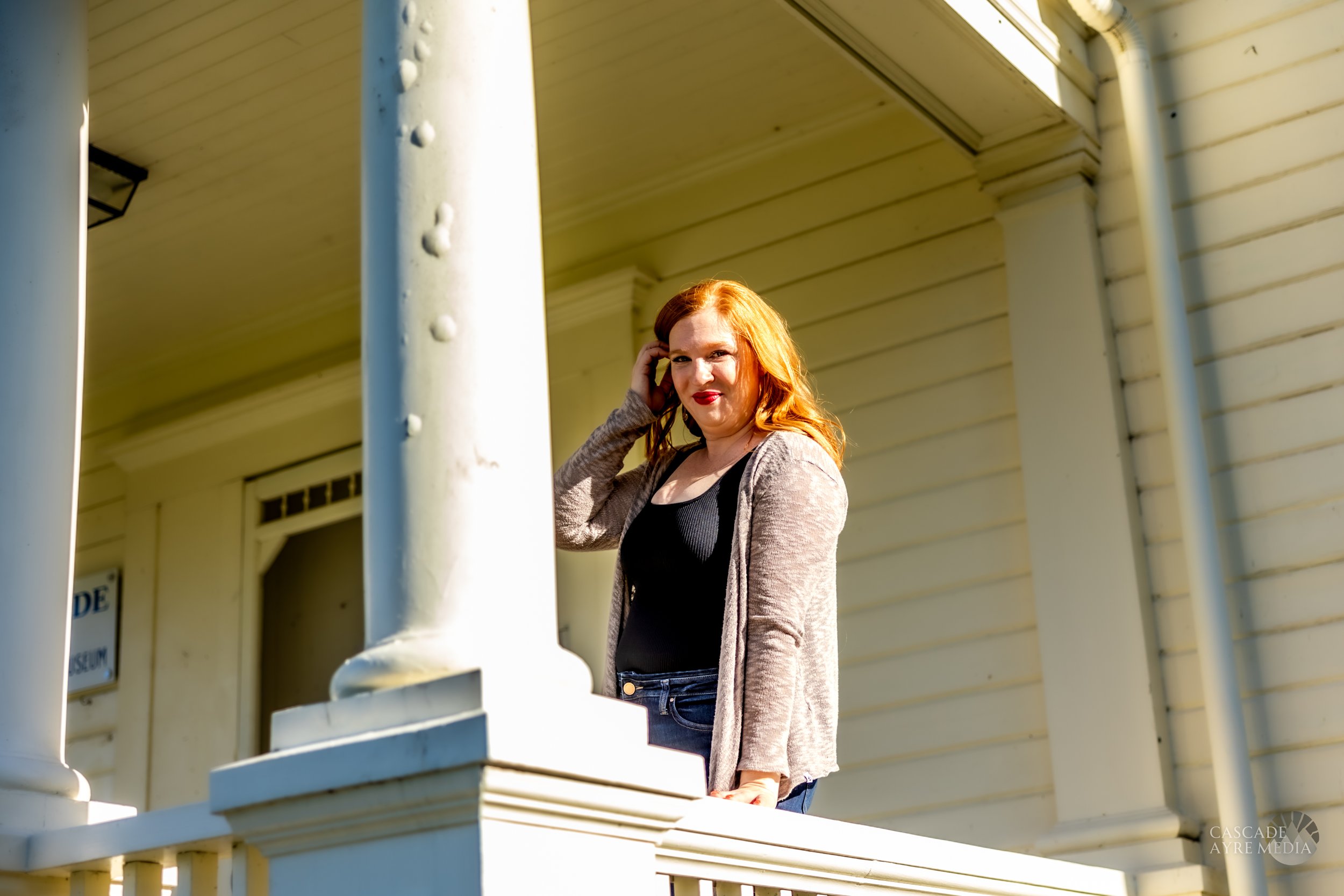 A person with red hair stands on a porch in front of a white building, touching their hair.