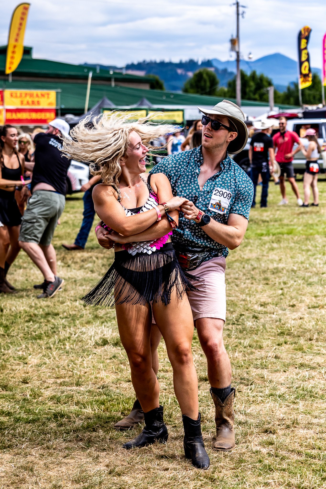 A man and woman dancing energetically at an outdoor event. The woman is wearing a fringed outfit and cowboy boots, while the man wears a hat, sunglasses, and shorts. Other attendees and food stalls are visible in the background.