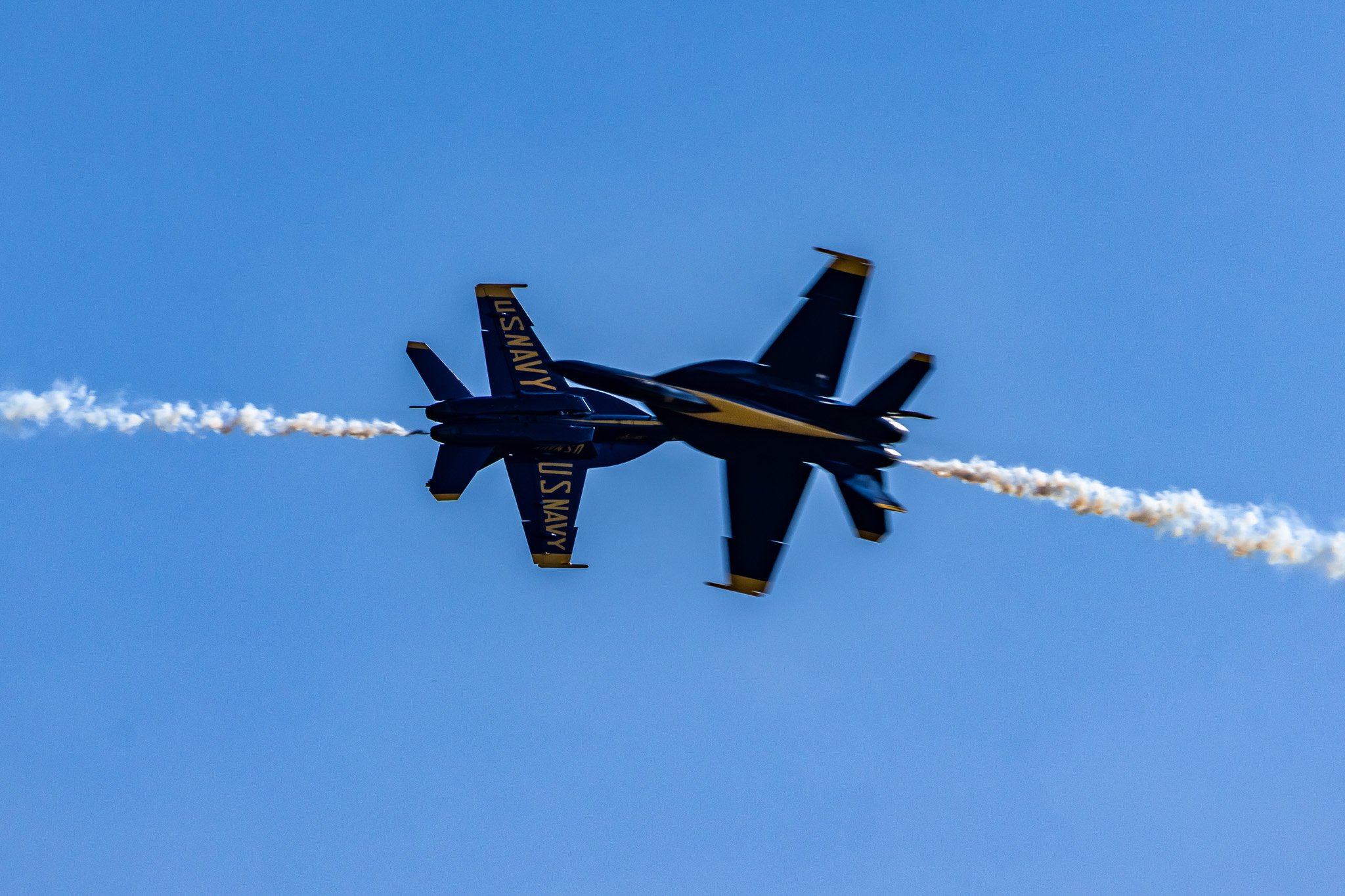 Two U.S. Navy Blue Angels jets performing an aerial maneuver with smoke trails against a clear blue sky.