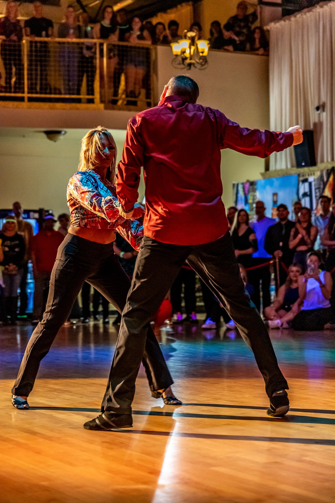 Two people dancing on a wooden floor with an audience watching in the background.