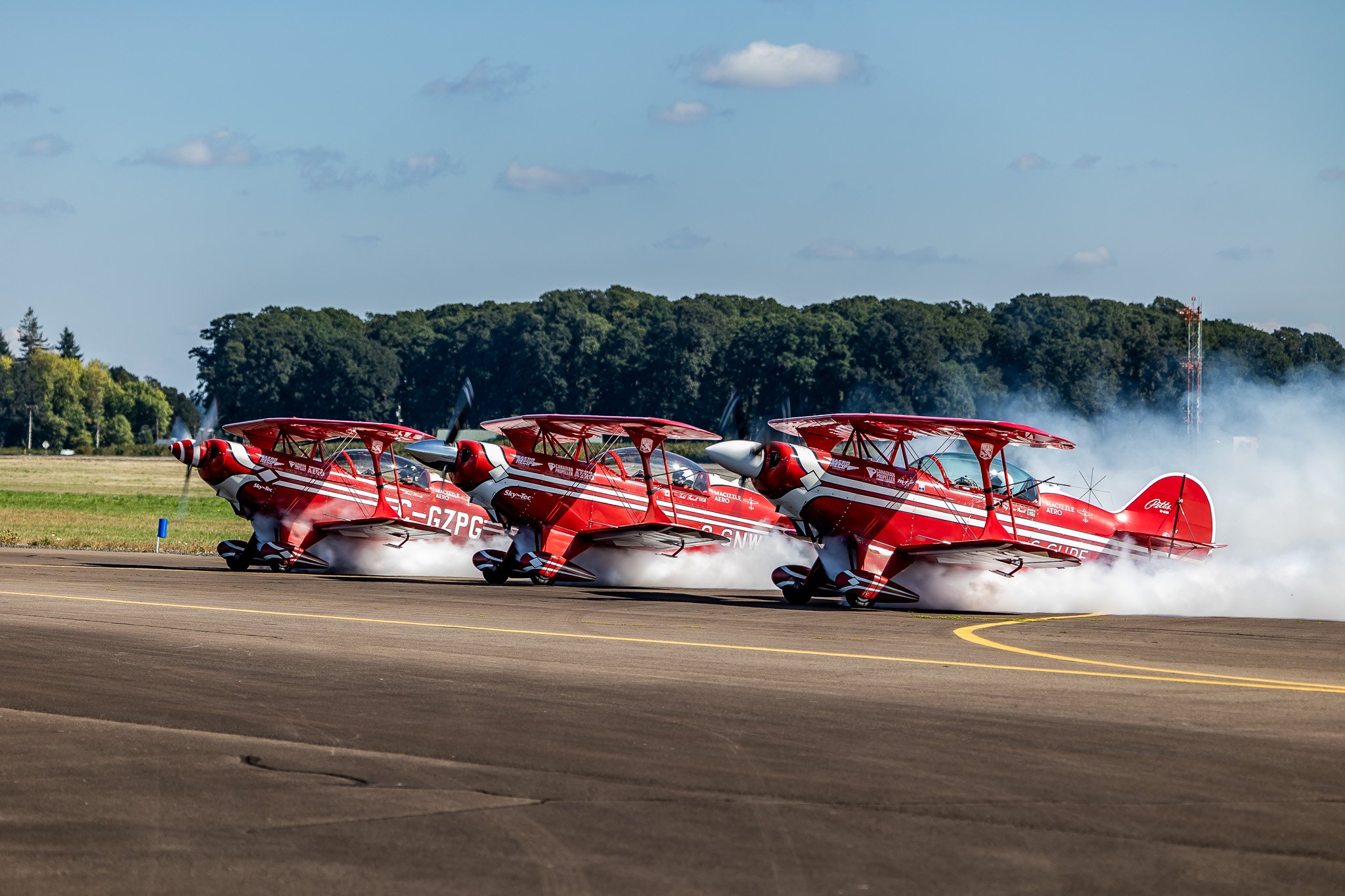 Three red stunt planes performing on a runway with smoke trailing behind, set against a backdrop of trees under a clear blue sky.