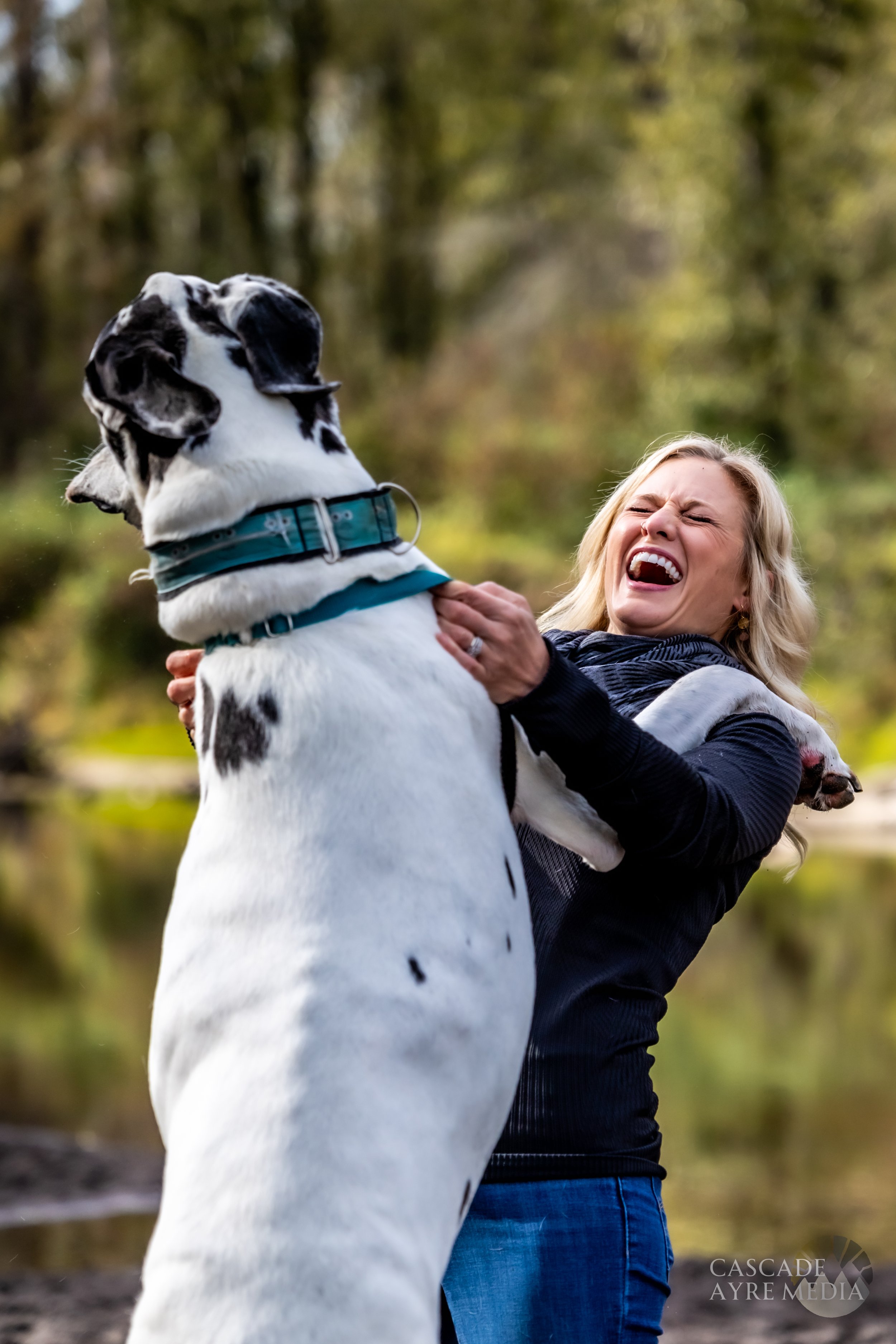 Woman laughing while a large spotted dog stands on its hind legs with its front paws on her shoulders in an outdoor setting.