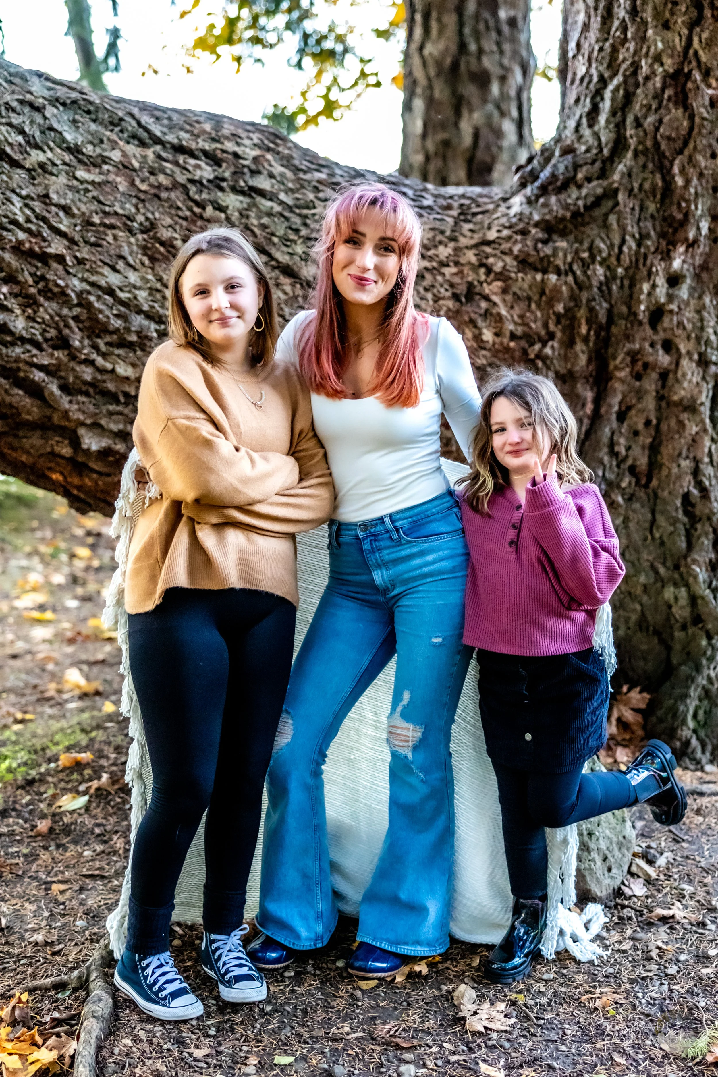 Three people posing in front of a large tree trunk outdoors, wearing casual clothing. The person in the middle is wearing a white top and blue jeans, standing with children on either side.
