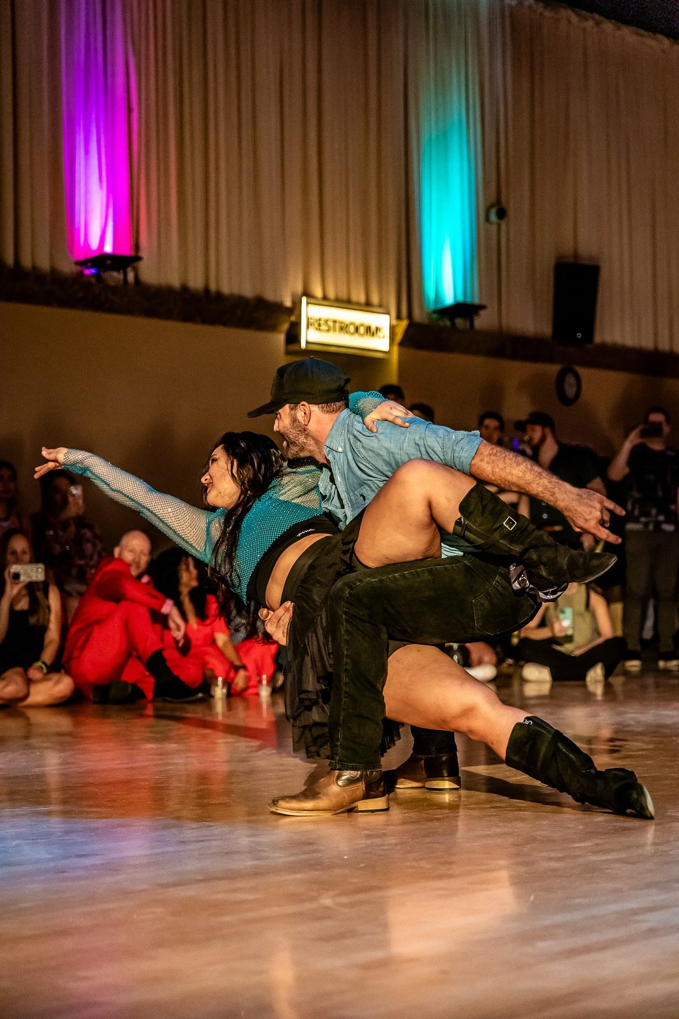 Couple dancing passionately on a dance floor, with onlookers seated and watching; the room is decorated with vibrant lighting.