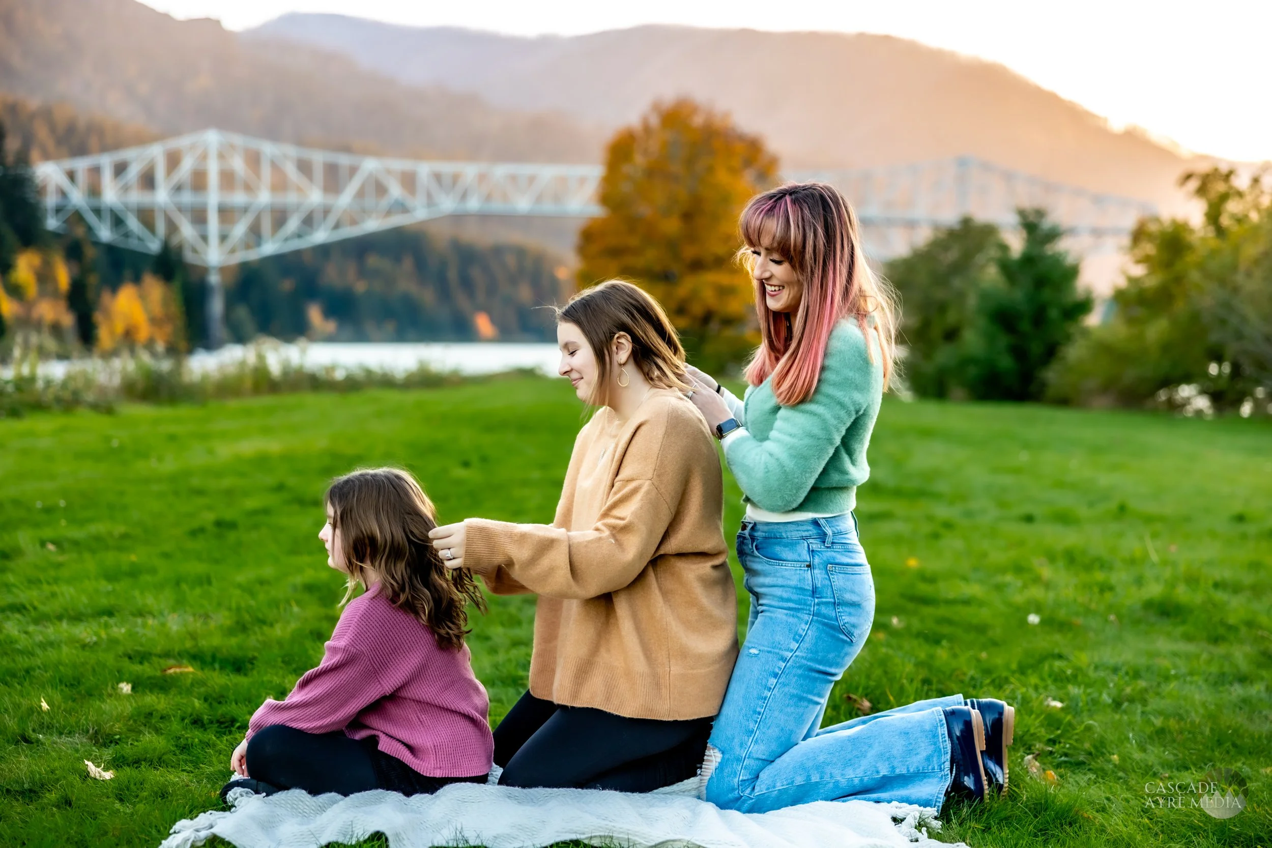 Three people sitting on grass in a scenic park, forming a braid chain with each other's hair; mountains, a bridge, and autumn foliage in the background.