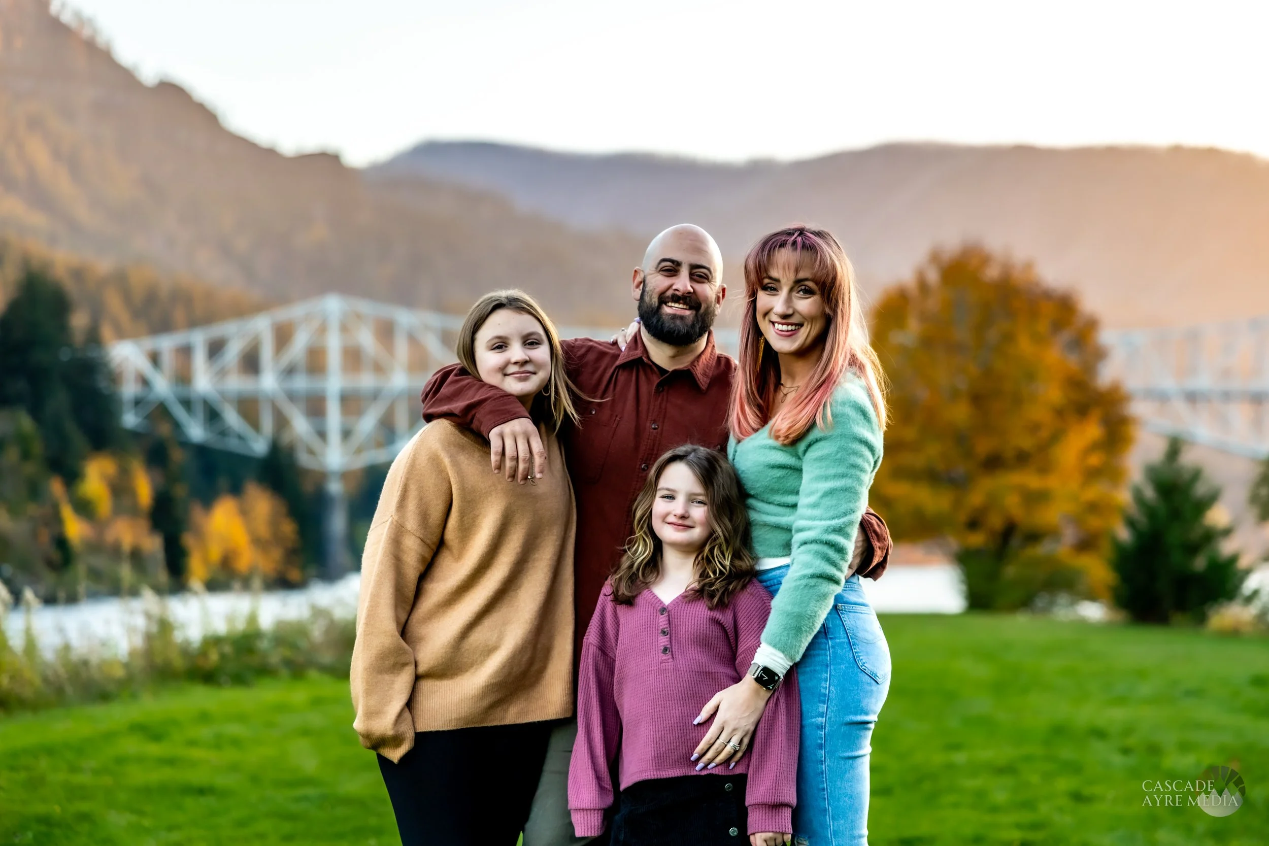 Four people smiling in a grassy area with autumn foliage and a bridge in the background.