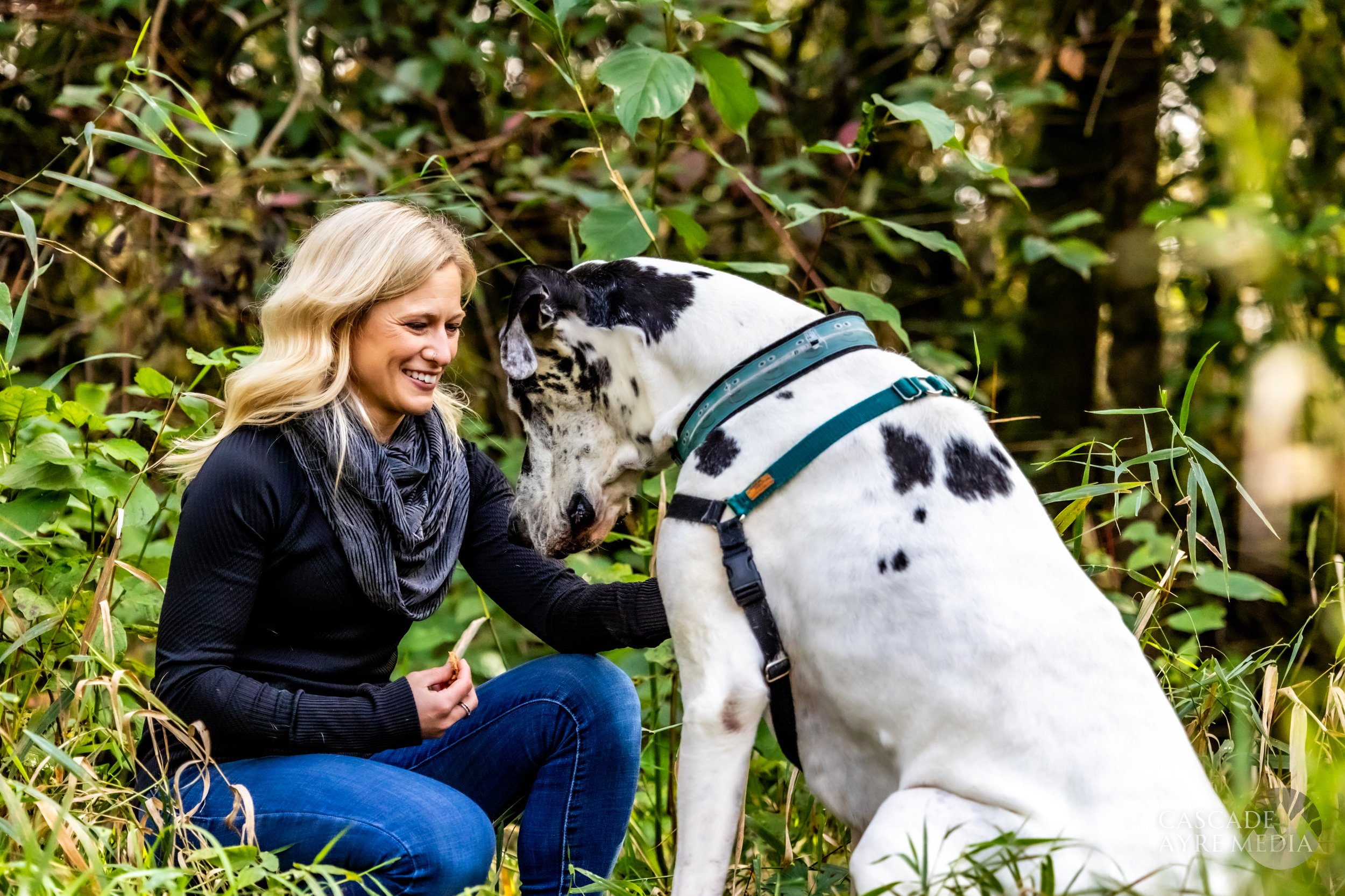 A woman sitting on the ground, smiling at a large black and white dog in a forest setting.