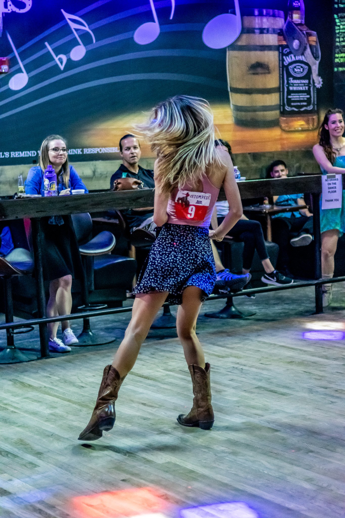 Woman dancing energetically in a bar, wearing cowboy boots and a floral skirt, with spectators watching.