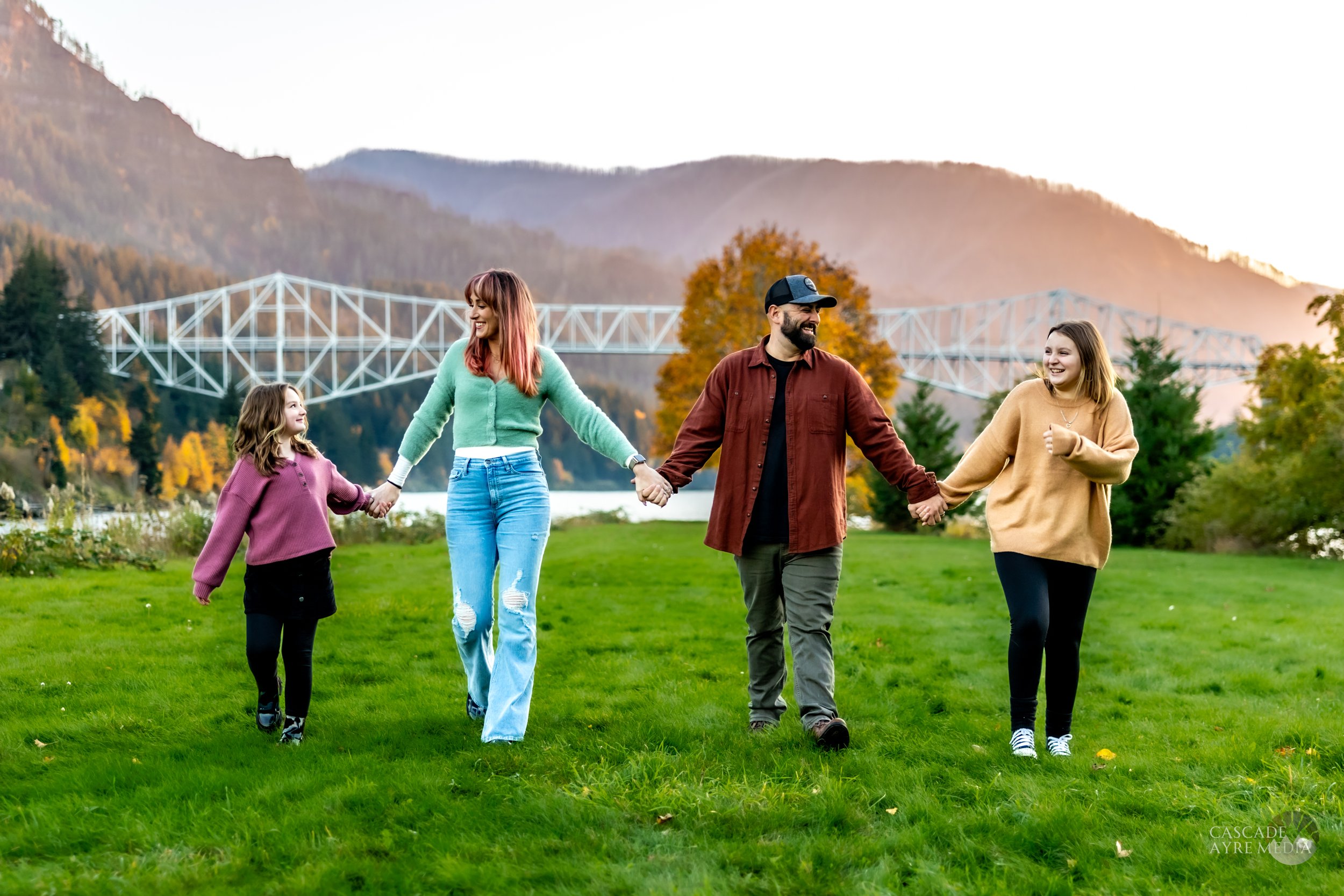 A family of four holding hands while walking on a grassy field with a scenic view of a bridge and mountains in the background.