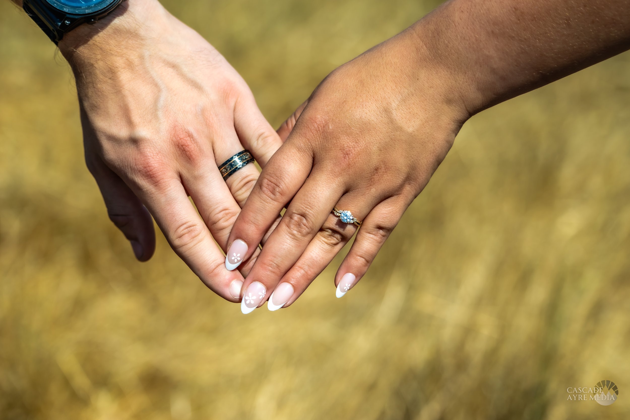 Two hands with rings, one wearing an engagement ring and the other a band, clasped together with a blurred outdoor background.
