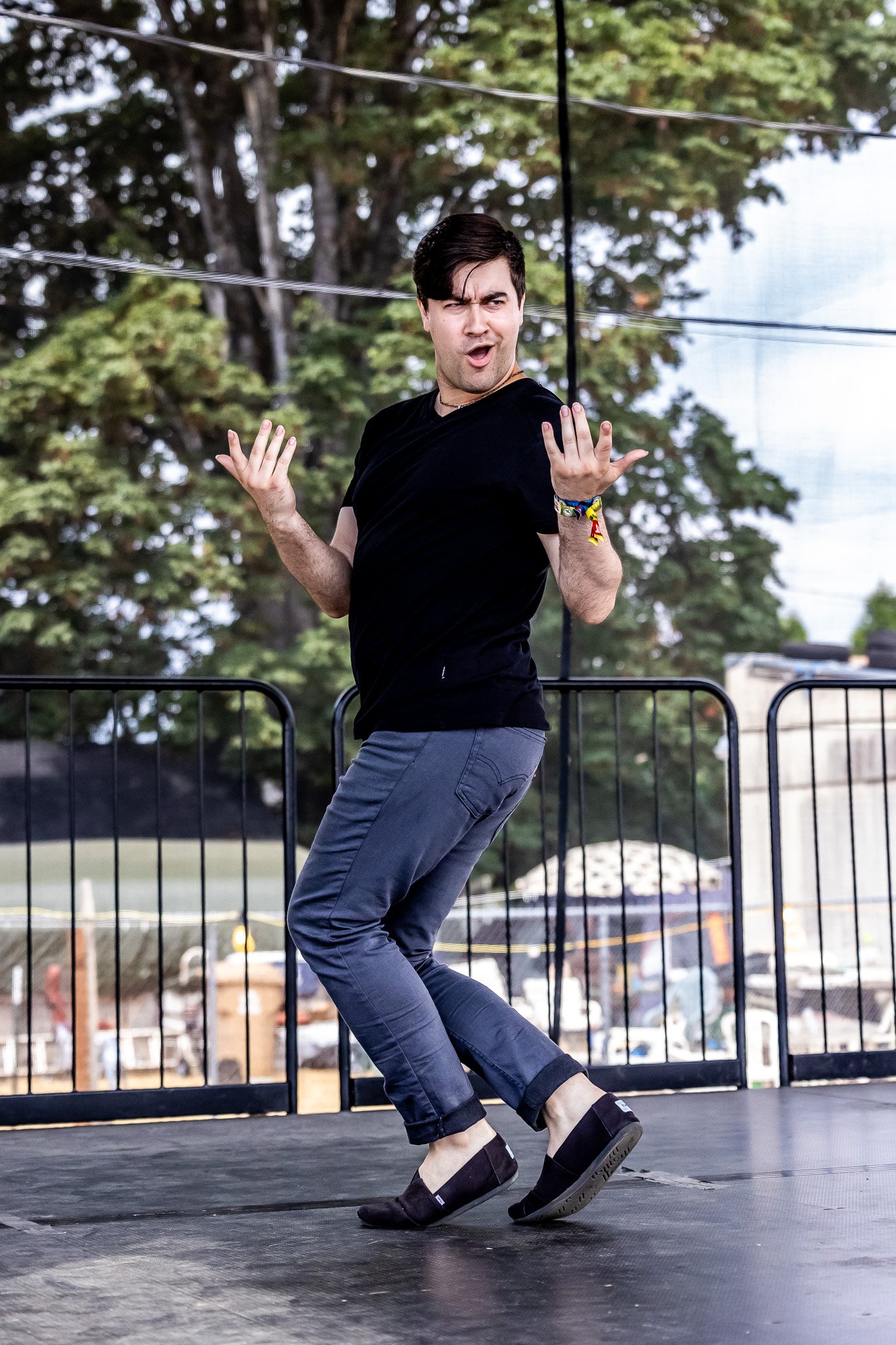 Person in black shirt and gray pants dancing energetically on stage with trees in background.