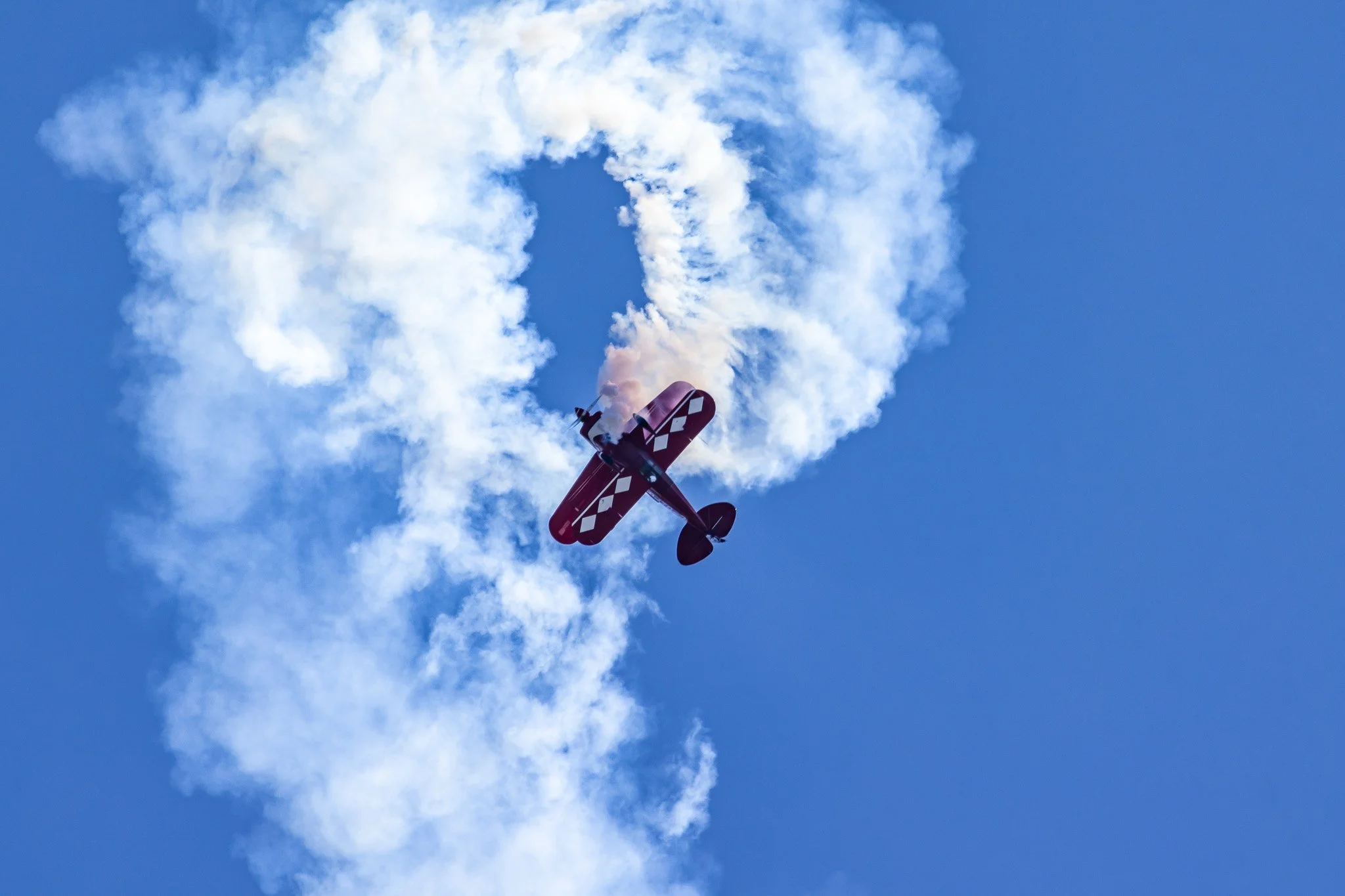 Red biplane performing aerobatics, leaving a smoke trail against a clear blue sky.