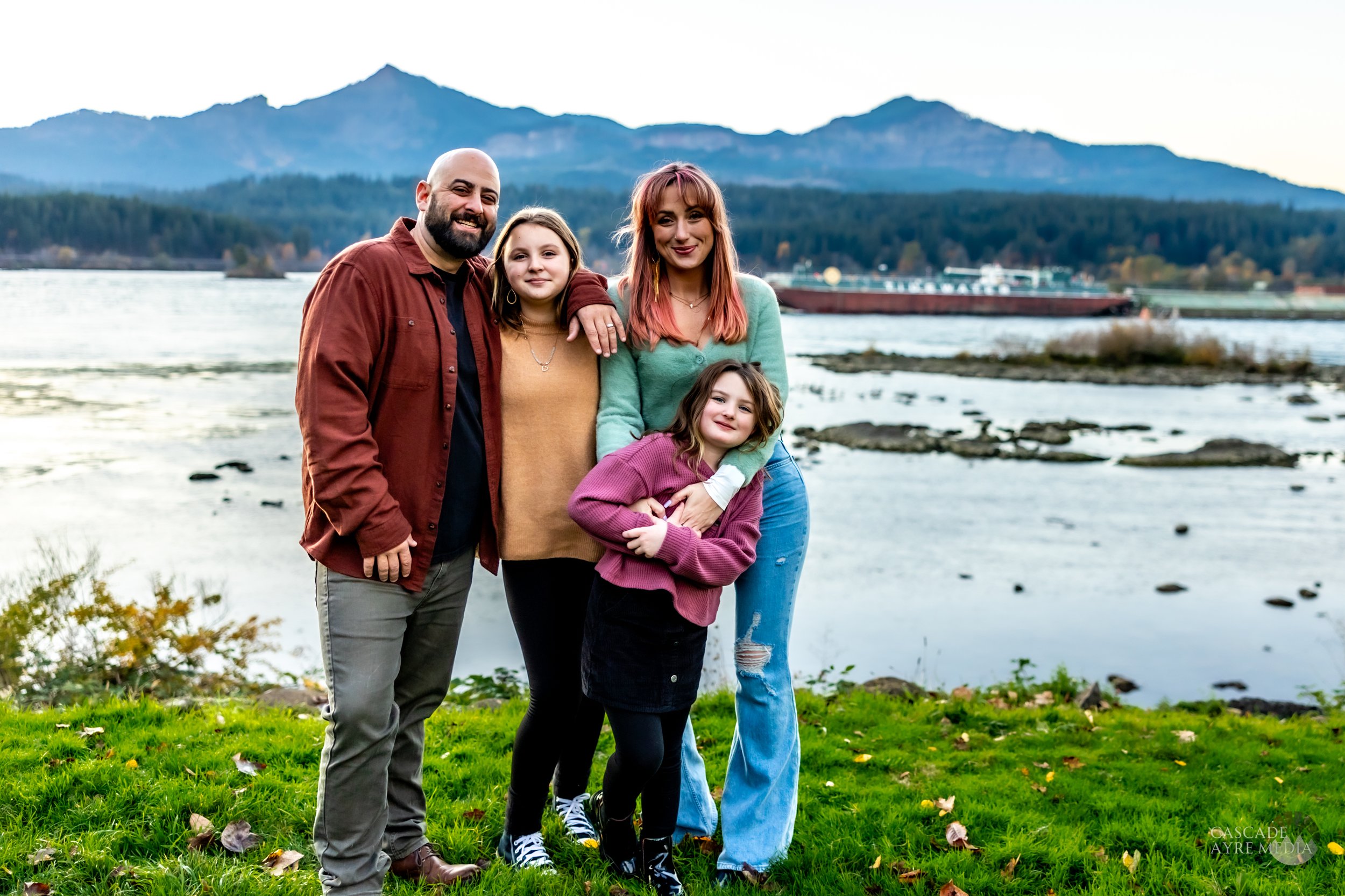 A family of four poses outdoors by a river with mountains in the background. The father is wearing an orange jacket, the mother a green sweater, one child in a brown sweater, and the other child in a pink sweater. They are standing on a grassy area.