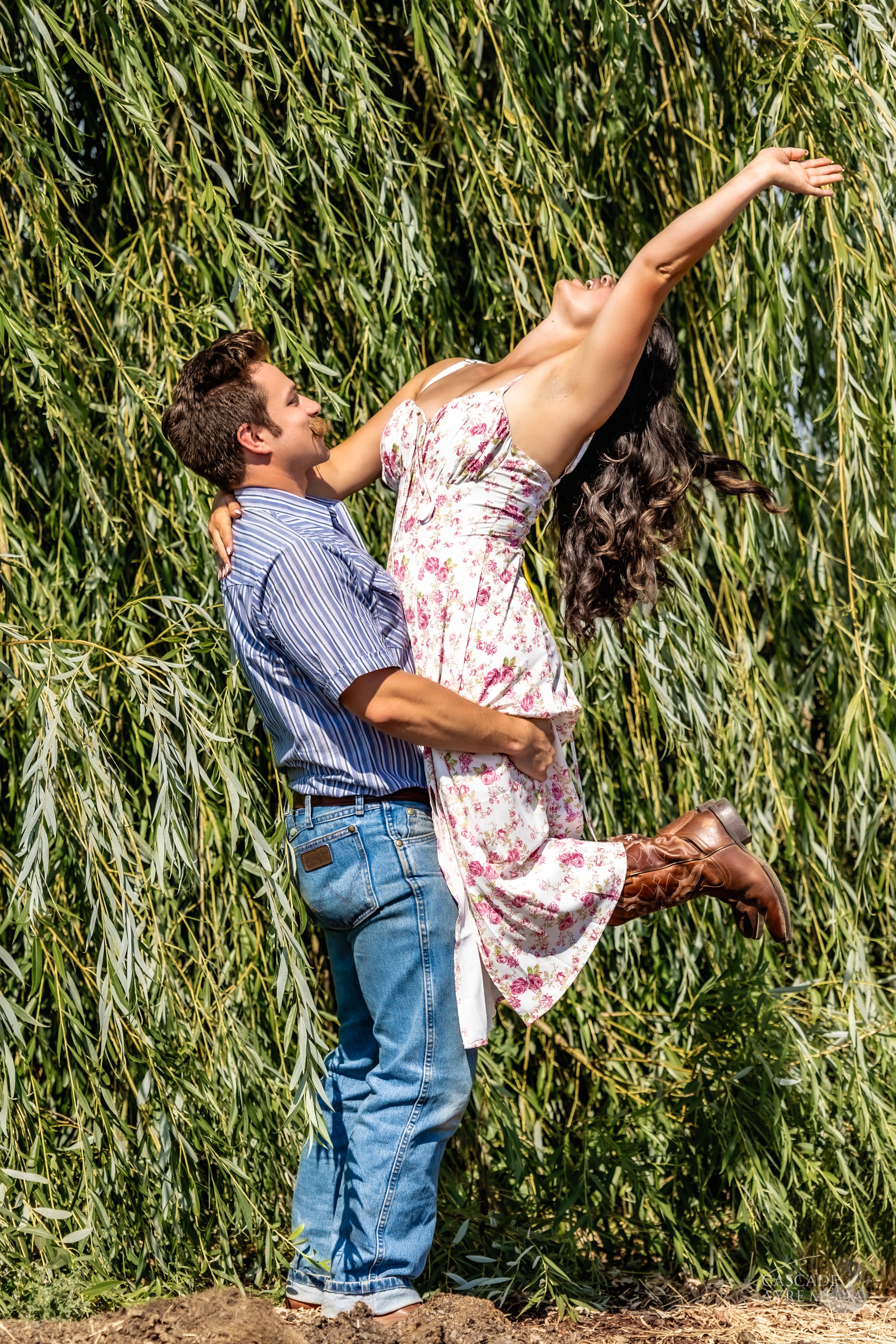A man lifts a woman in a floral dress and cowboy boots in front of a background of green willow trees. The woman appears joyful, with her arms raised.