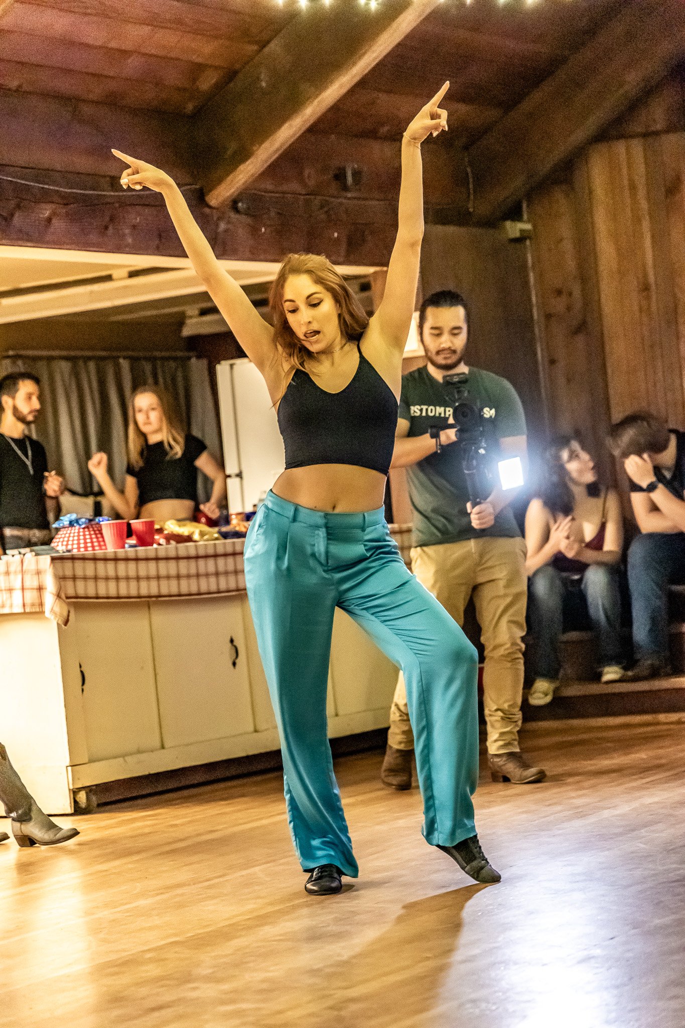 A woman dancing with arms raised, wearing a black top and blue pants, surrounded by people and a photographer holding a camera with a flash inside a wooden room.