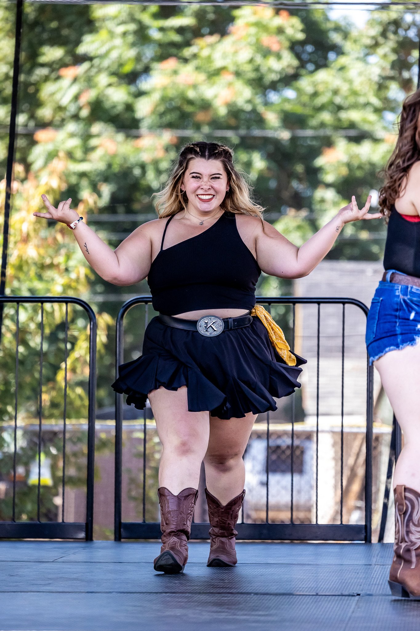 Woman in black top dancing on stage outdoors, wearing a black skirt and brown cowboy boots.