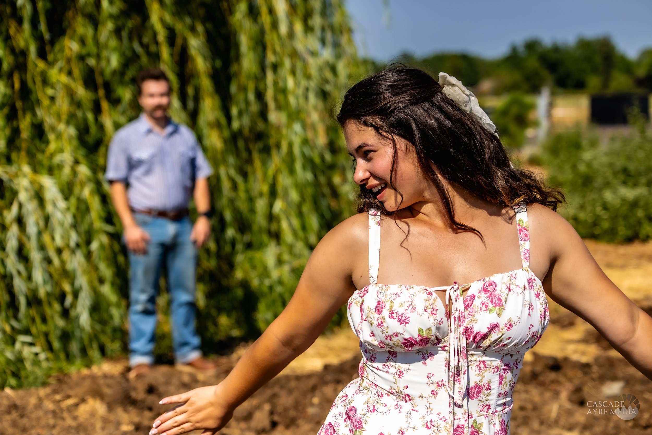 A woman in a floral dress smiling outside with a blurred man in the background near greenery.