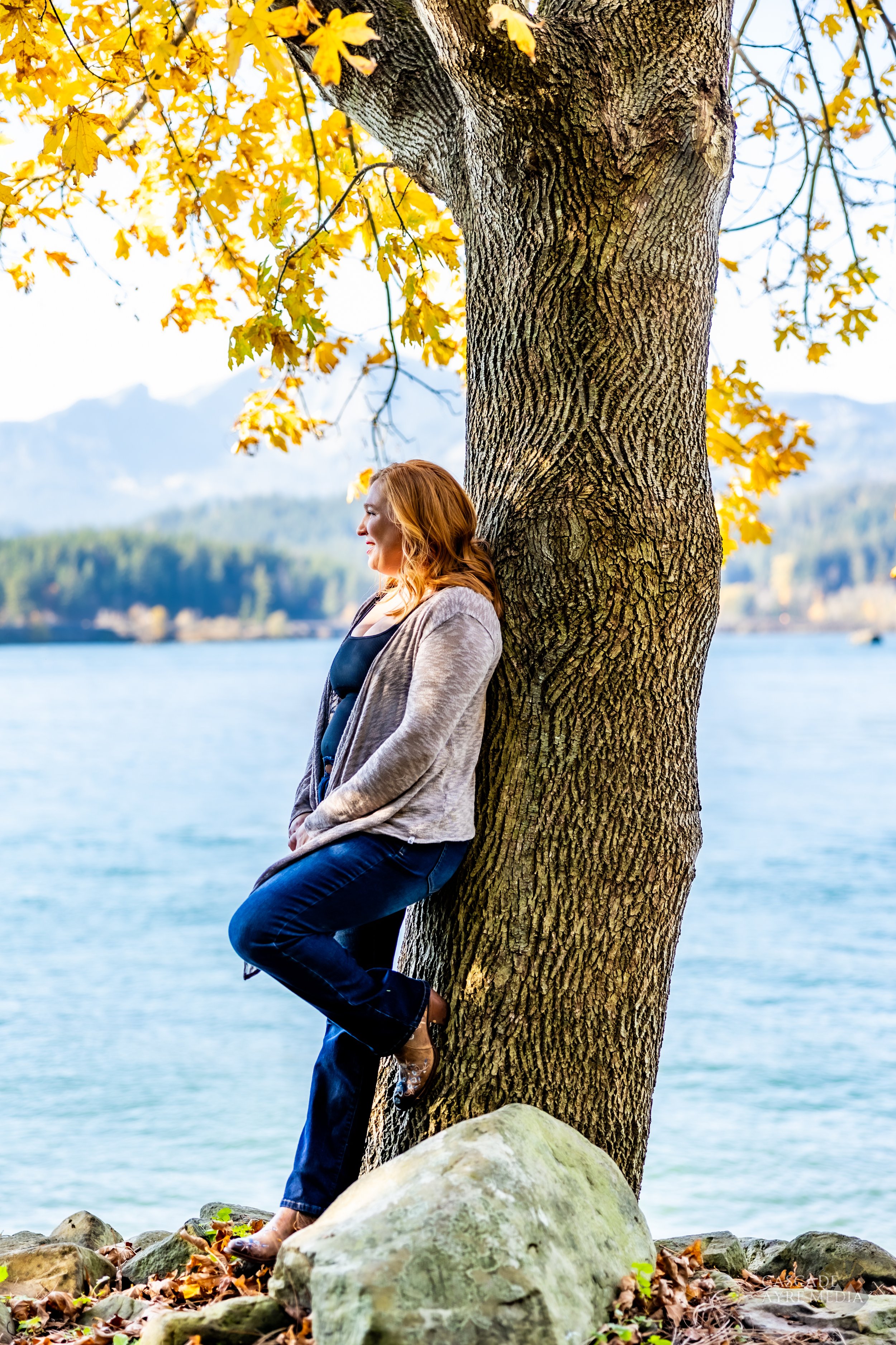 A woman with red hair leans against a tree beside a lake with mountains in the background and autumn leaves on the tree.