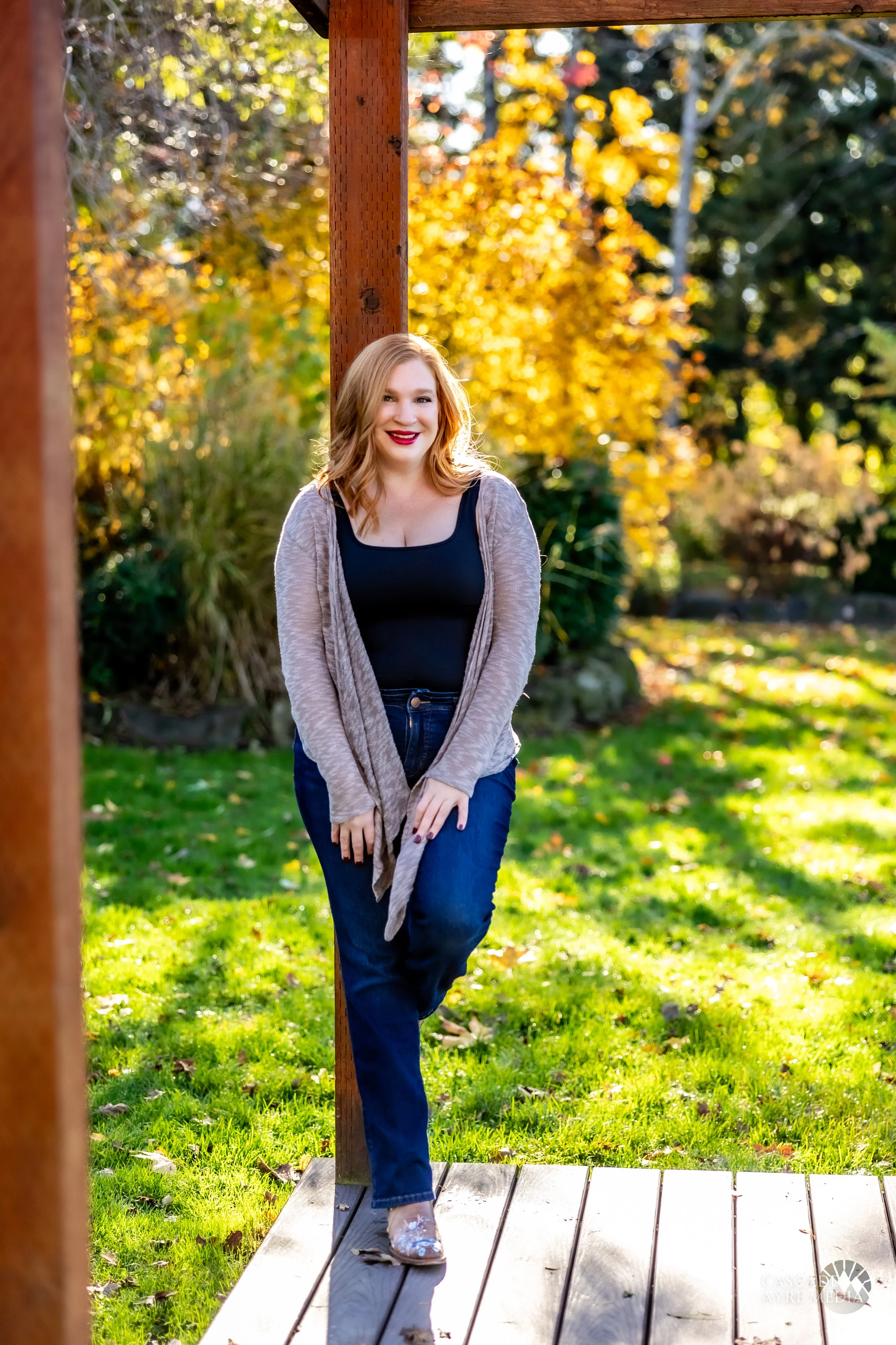 Woman with long red hair wearing a black top and light cardigan, standing on a wooden platform in a park with autumn foliage.