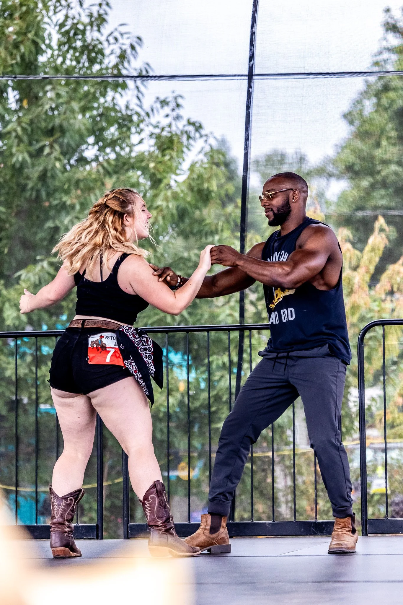 Two people dancing energetically on an outdoor stage, one wearing a black top and shorts with cowboy boots, the other in a sleeveless shirt and pants with brown boots, surrounded by green trees.