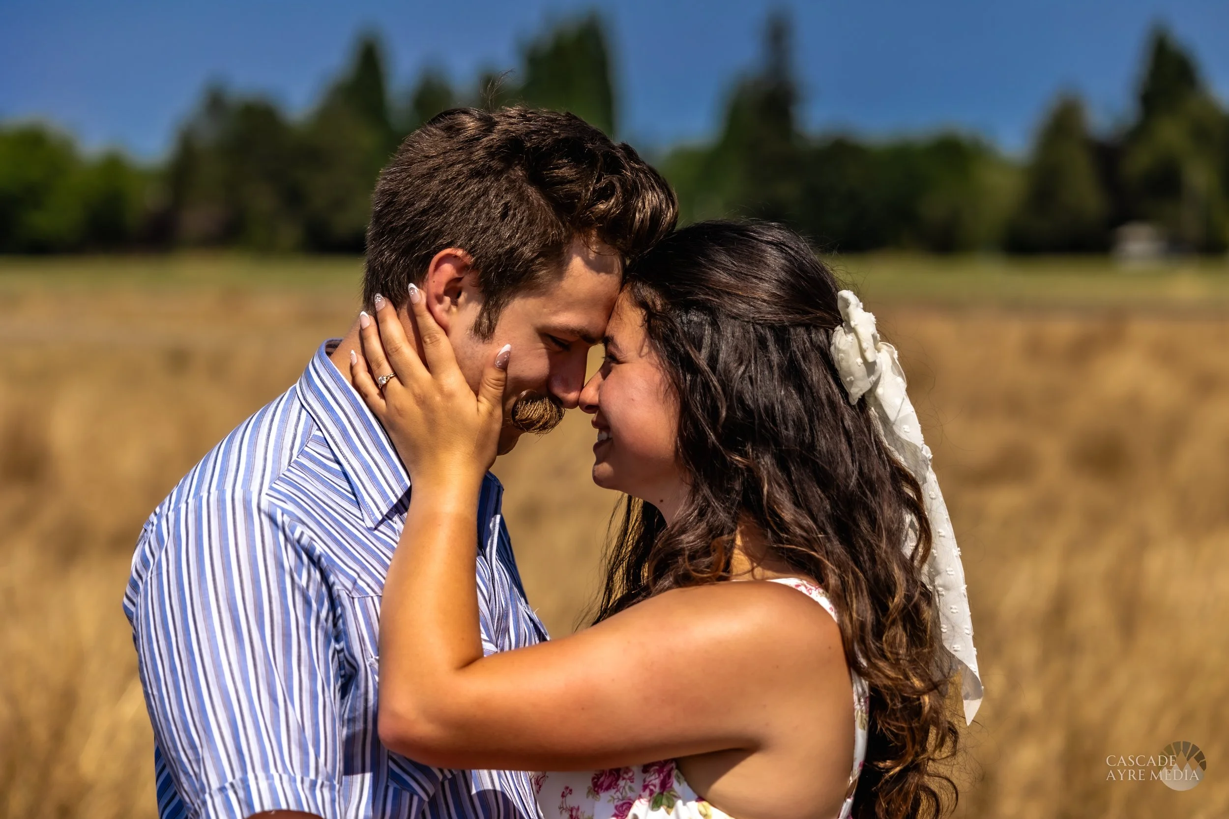 A couple embracing and smiling in a field under a clear blue sky.