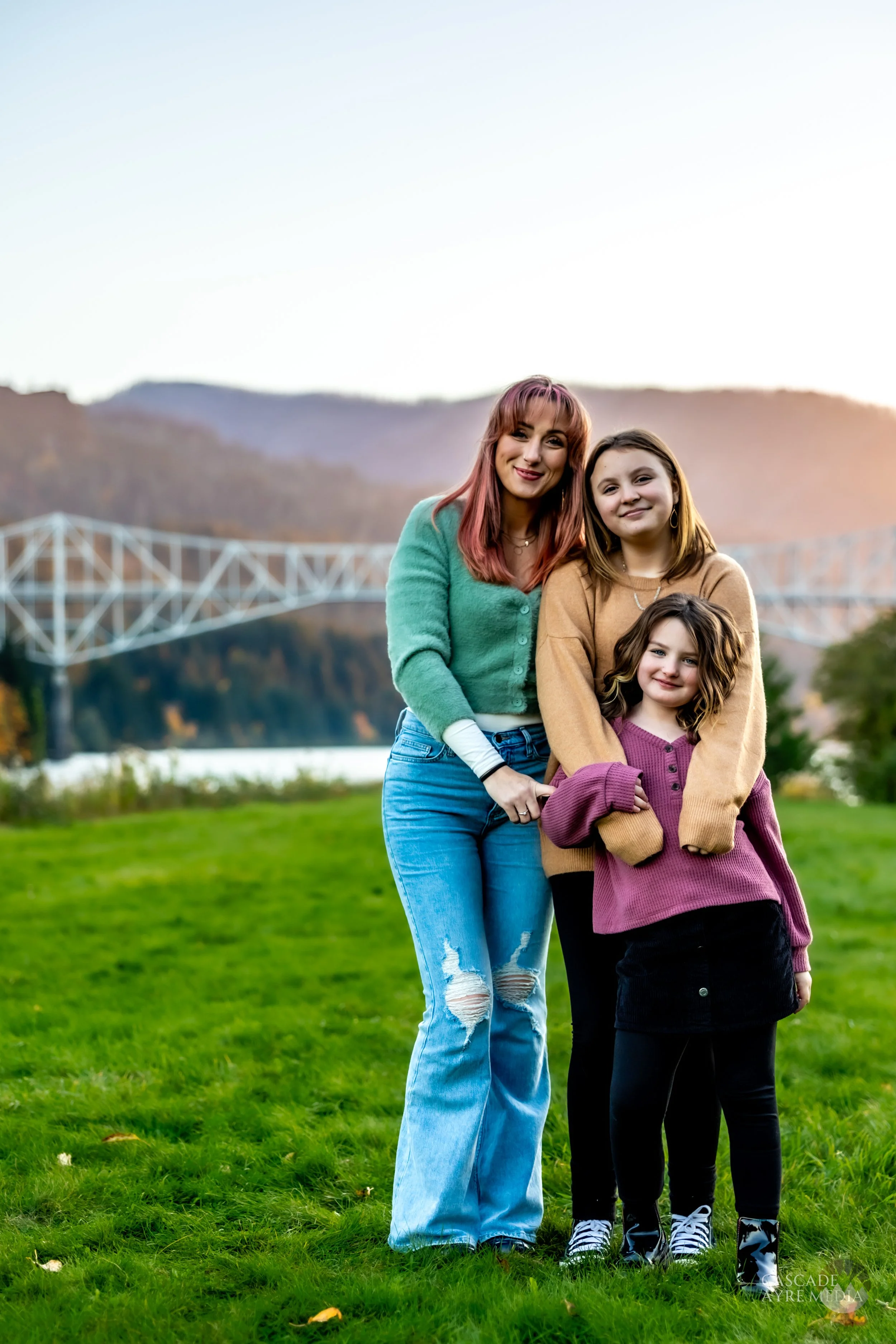 Three people standing on grass with a bridge and hills in the background, wearing casual outfits.