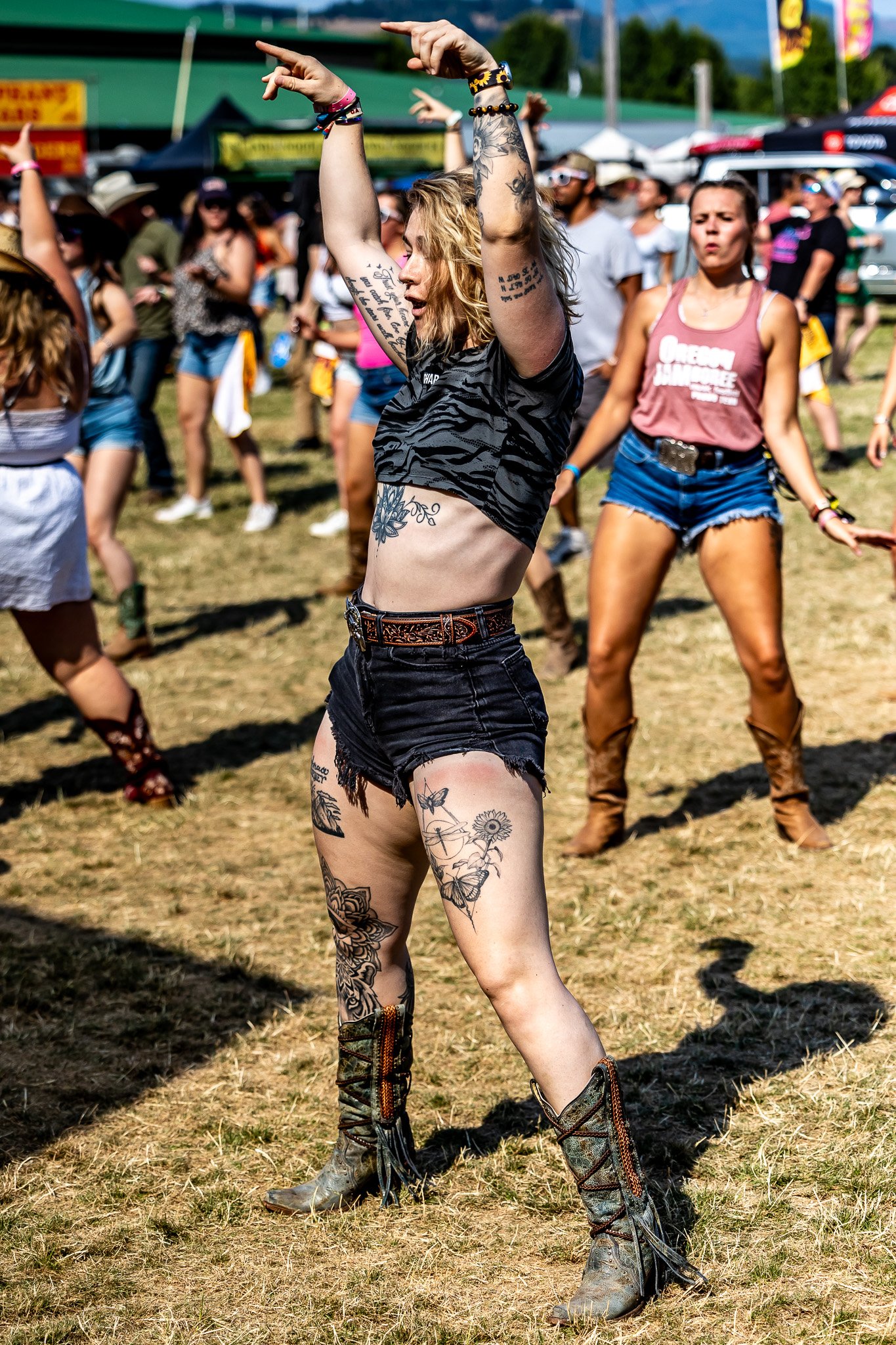 People dancing at an outdoor festival with tattoos and casual clothing, including cowboy boots. Some are wearing tank tops and shorts, enjoying the sunny day.