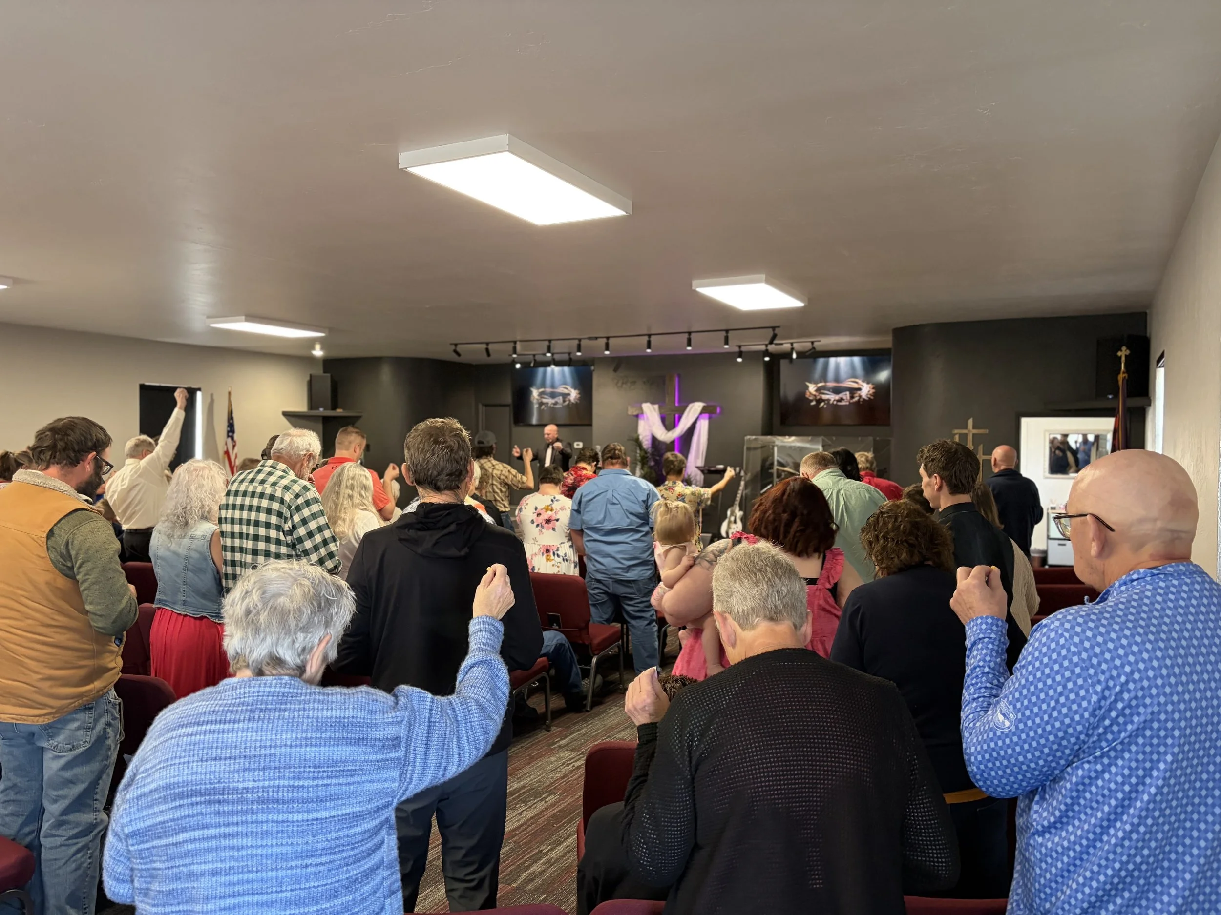 A congregation inside a church or chapel, standing and praying with raised hands, facing the altar and cross at the front, during a religious service.