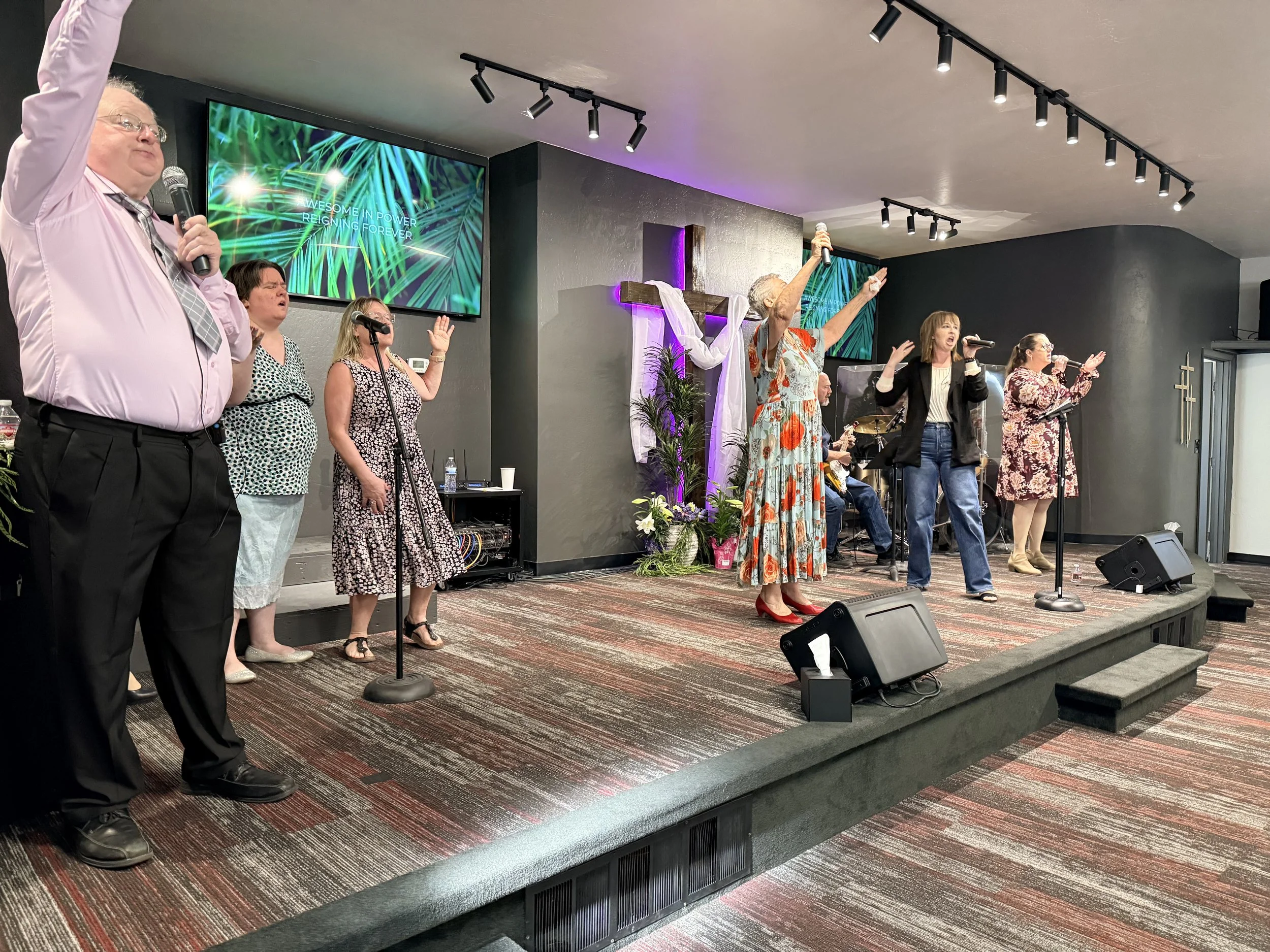 A group of diverse people singing and worshipping on stage in a church with a cross and a large screen displaying green leaves and the words 'Awesome in Power Reigning Forever'.