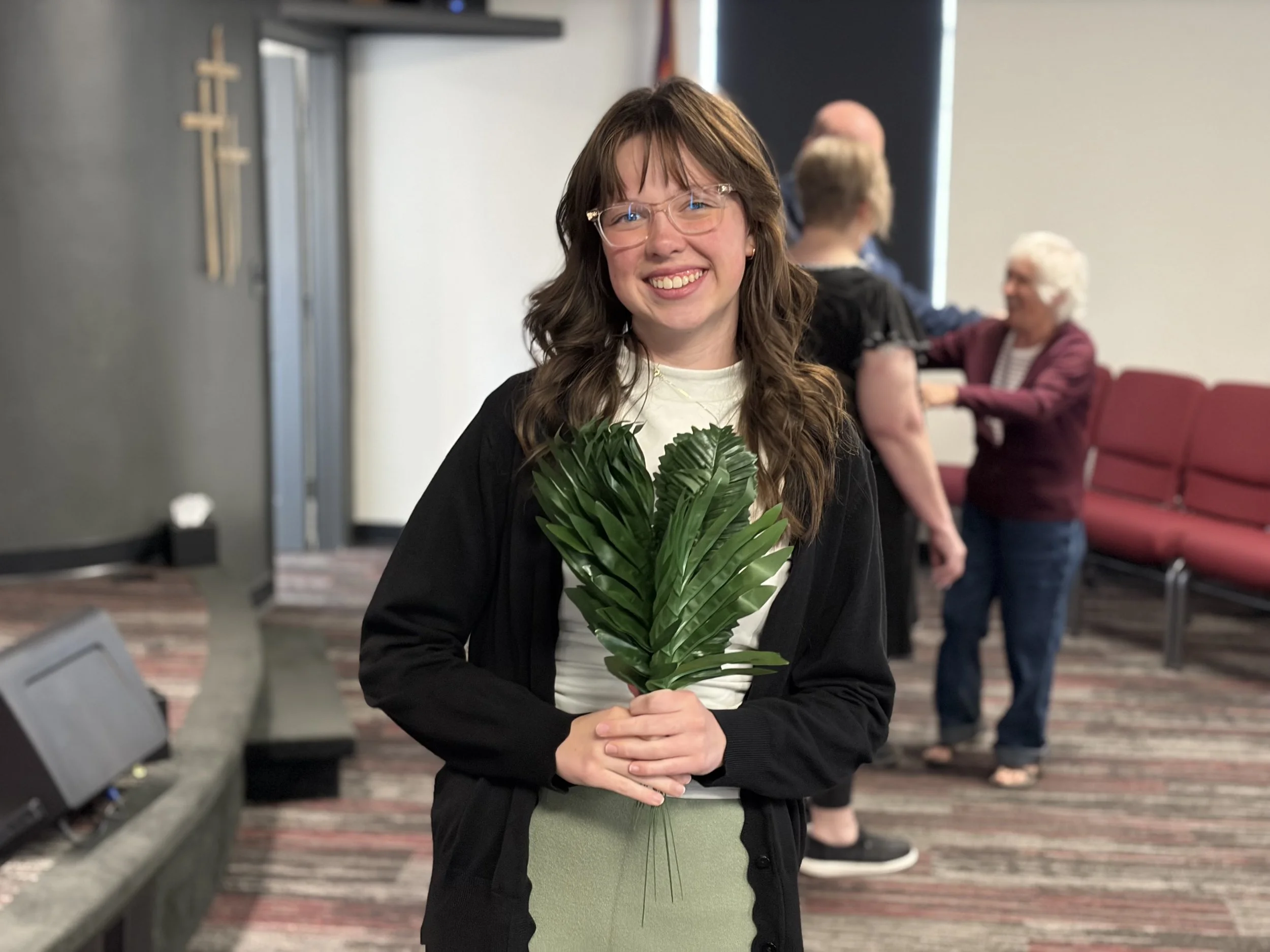 A young woman with long wavy brown hair and glasses smiling and holding a large green leaf plant in a room with red chairs and people in the background.