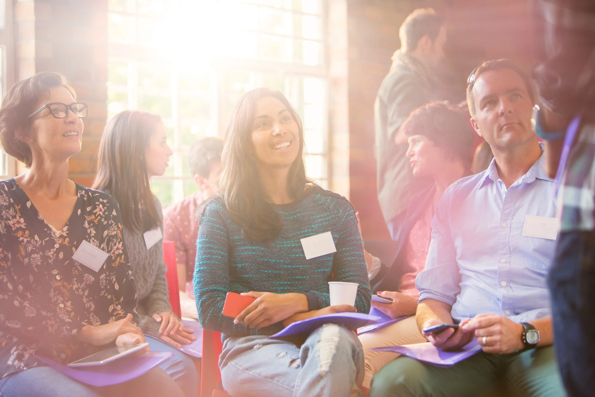 Group of diverse people attending a workshop or seminar, sitting on chairs with notebooks and papers, some smiling and conversing, in a well-lit room with large windows.