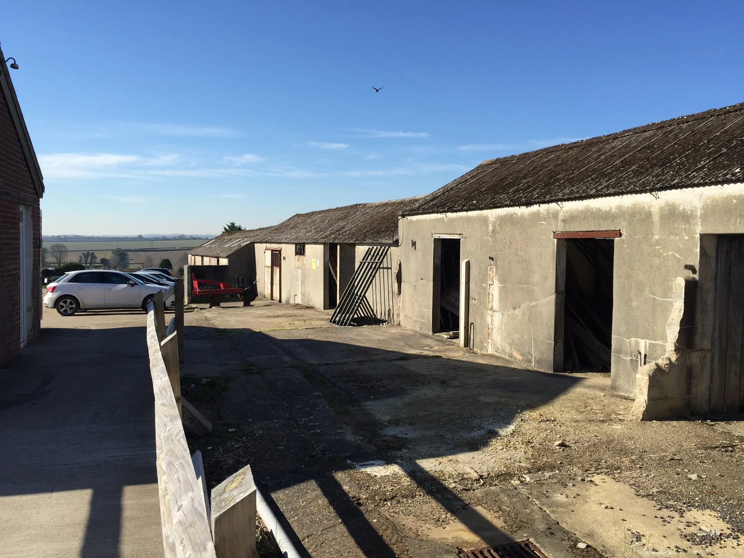 A row of old, weathered garages with open doors under a clear blue sky, some with damaged walls and leaning gates, with parked cars and a landscape visible in the background.