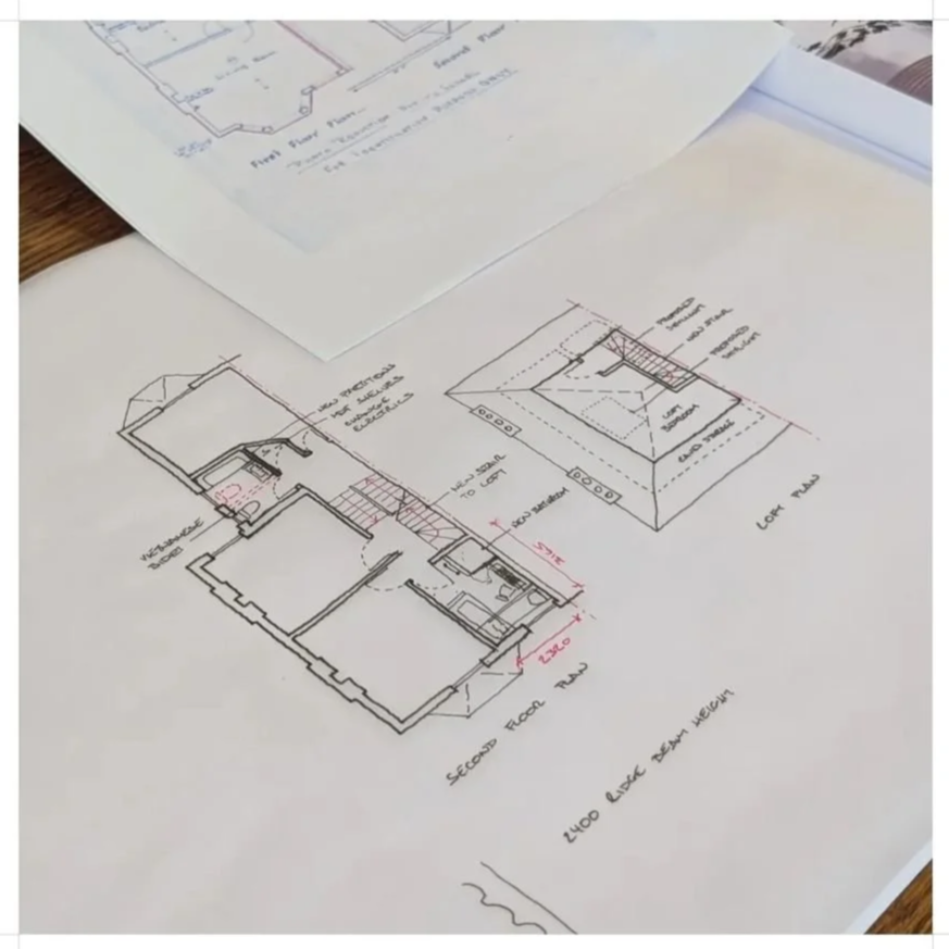 Architectural blueprints and floor plans of a house on white paper, including a second-floor plan and roof design, laid out on a wooden table.