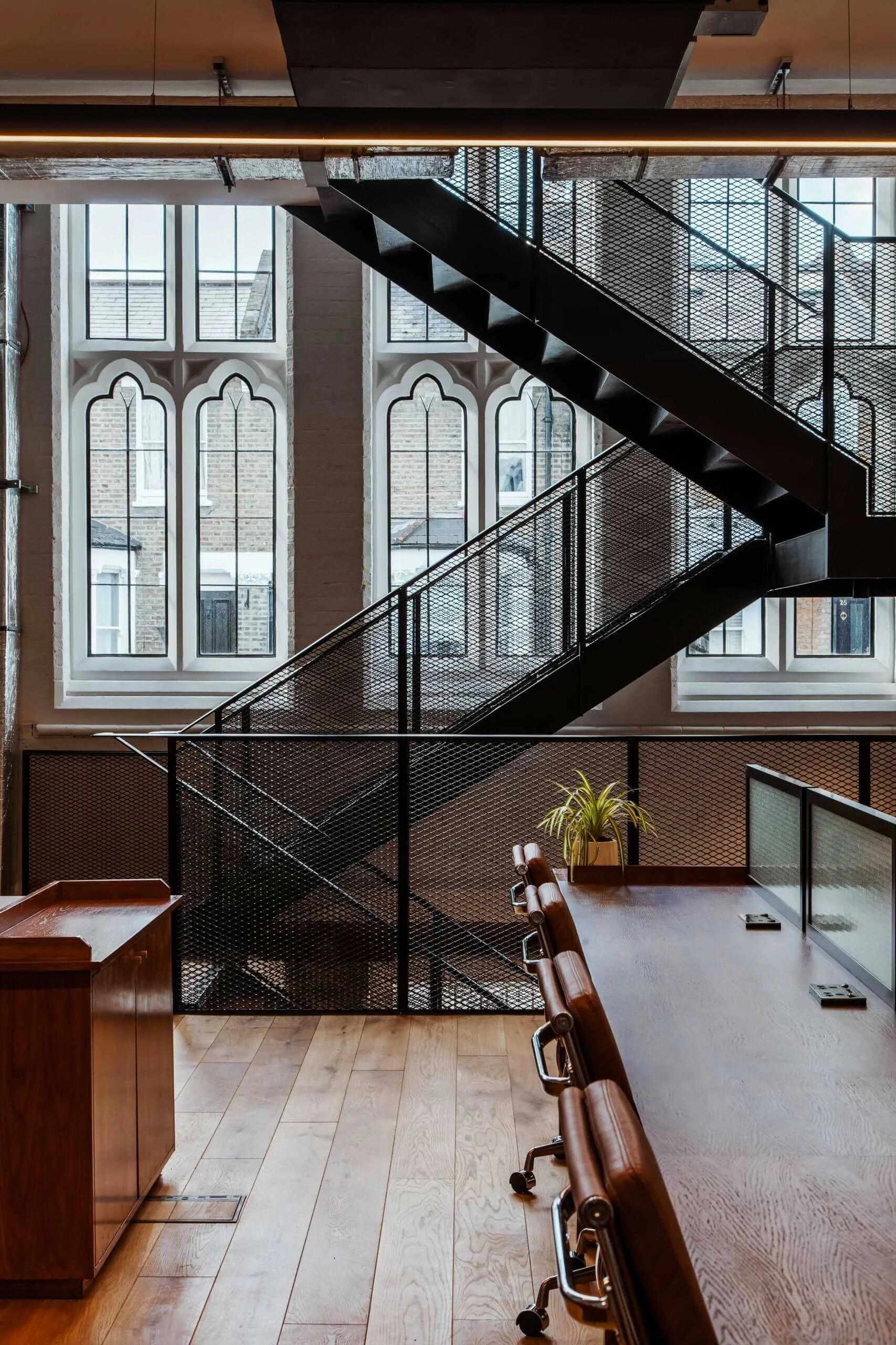 Interior of an office with wooden floor, row of brown chairs, a desk, a black metal staircase, and large gothic-style windows.