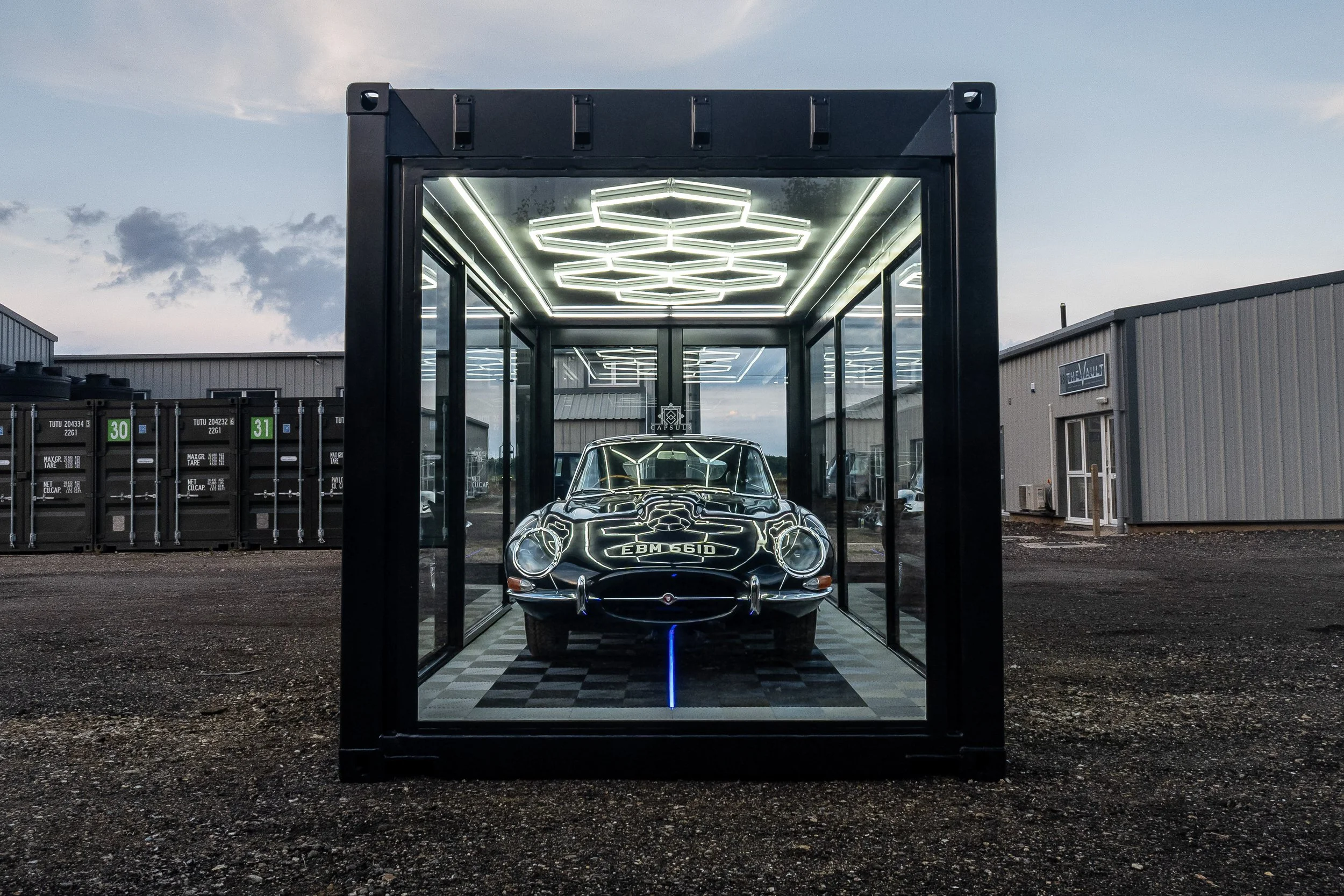 A vintage black sports car displayed inside a glass enclosure with neon lighting reflections, set outdoors during dusk.