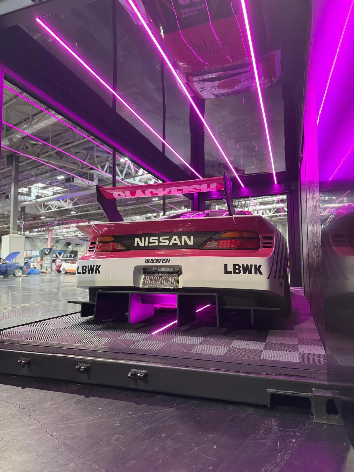 A Nissan race car displayed indoors with purple neon lighting reflecting on its surface, featuring a large rear spoiler and a custom license plate that reads 'HOOMIGAN'.