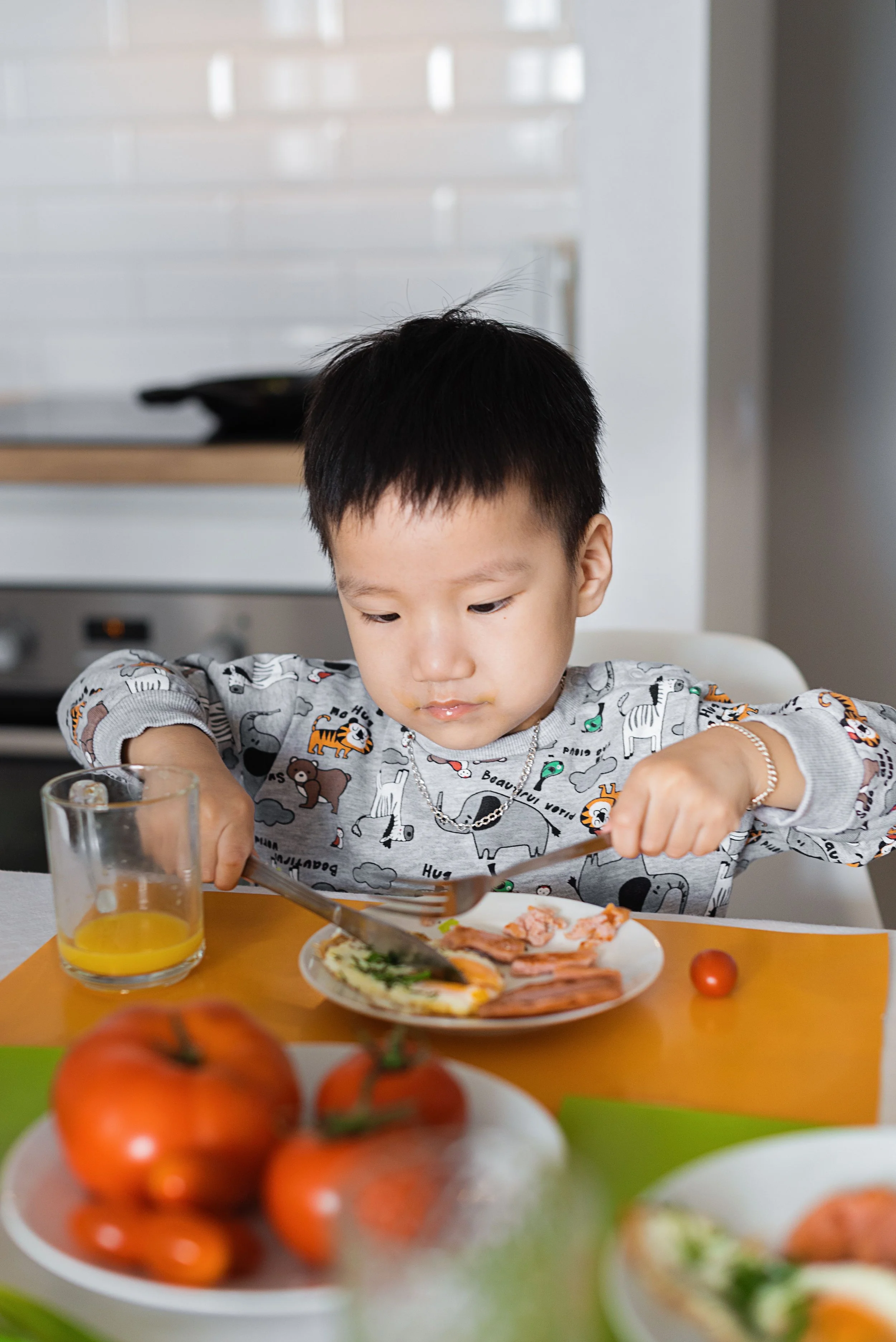 A young boy with short black hair eating breakfast at a table. There is a glass of orange juice and a plate of food including an egg, bacon, and toast in front of him. The background shows part of a kitchen with a white backsplash.