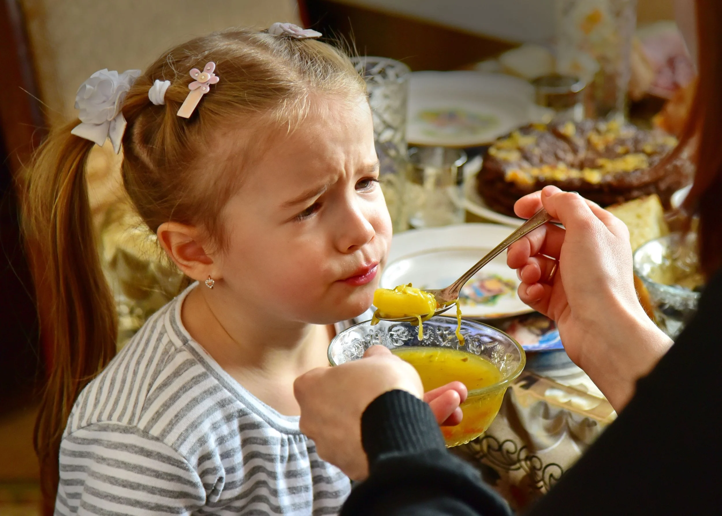 A young girl with blond hair, white bows, and a pink star-shaped hair clip looking at a spoonful of yellow food, likely soup, being held by an adult's hand. The background features a table set with local dishes, glassware, and decorative plates, suggesting a family meal or gathering.