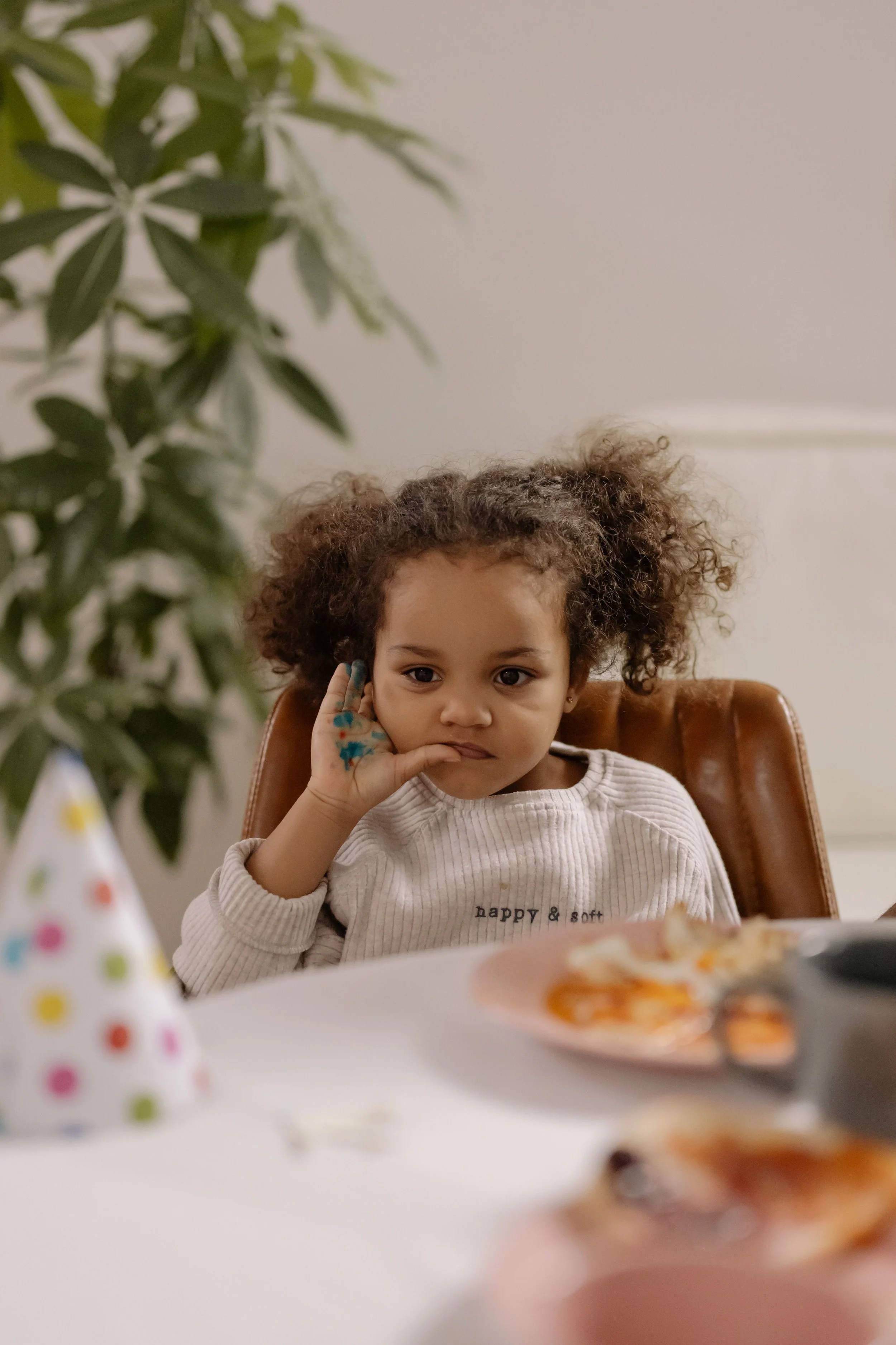 A young girl with curly hair sitting at a table, resting her hand on her face, wearing a sweater that says 'happy & soft'. There is a blurred party hat and some food on the table, and a large green plant is visible on the left side of the image.