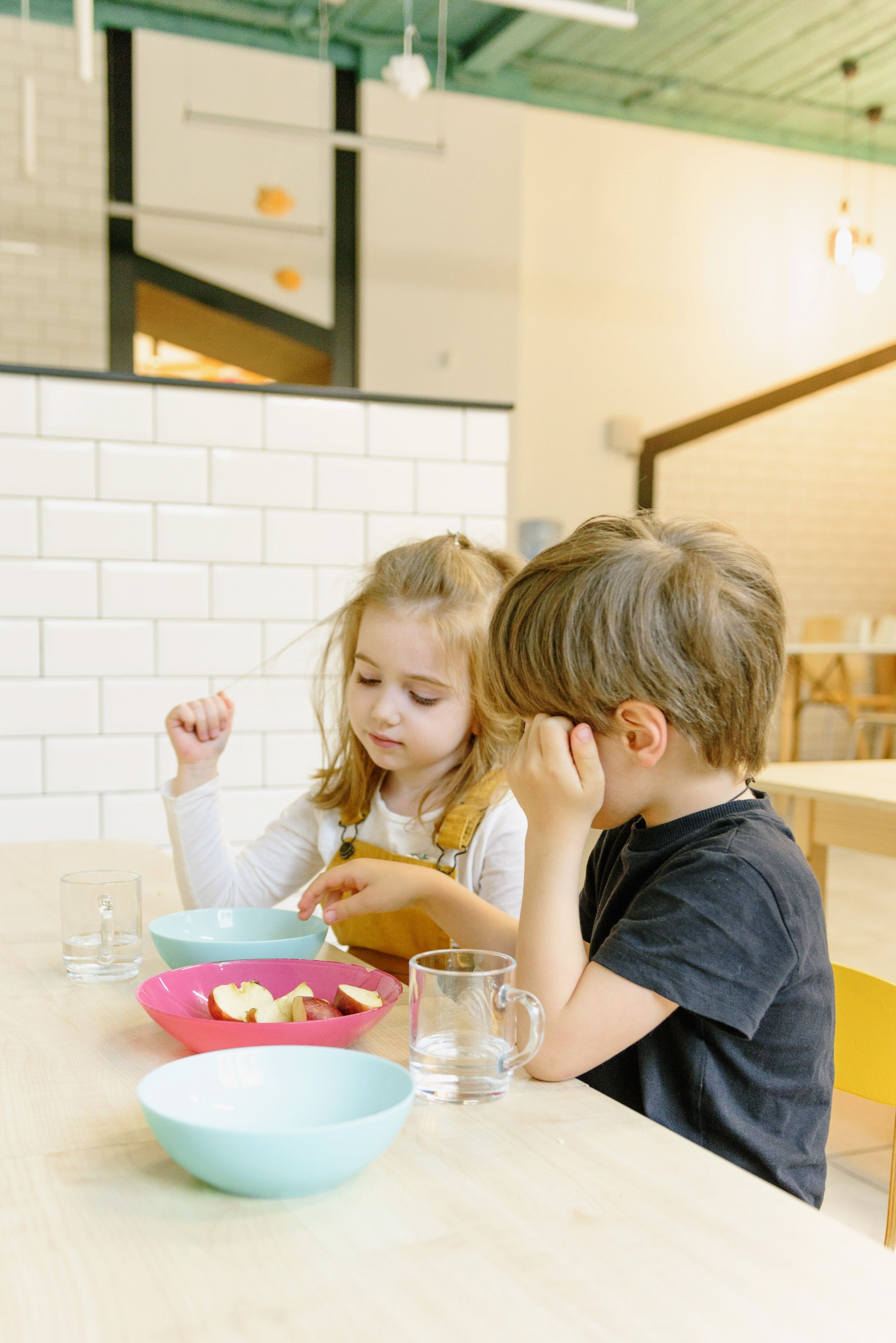 Two children sitting at a wooden table with bowls of apples and glasses of water, in a brightly lit, modern indoor setting with white brick walls and a green ceiling.