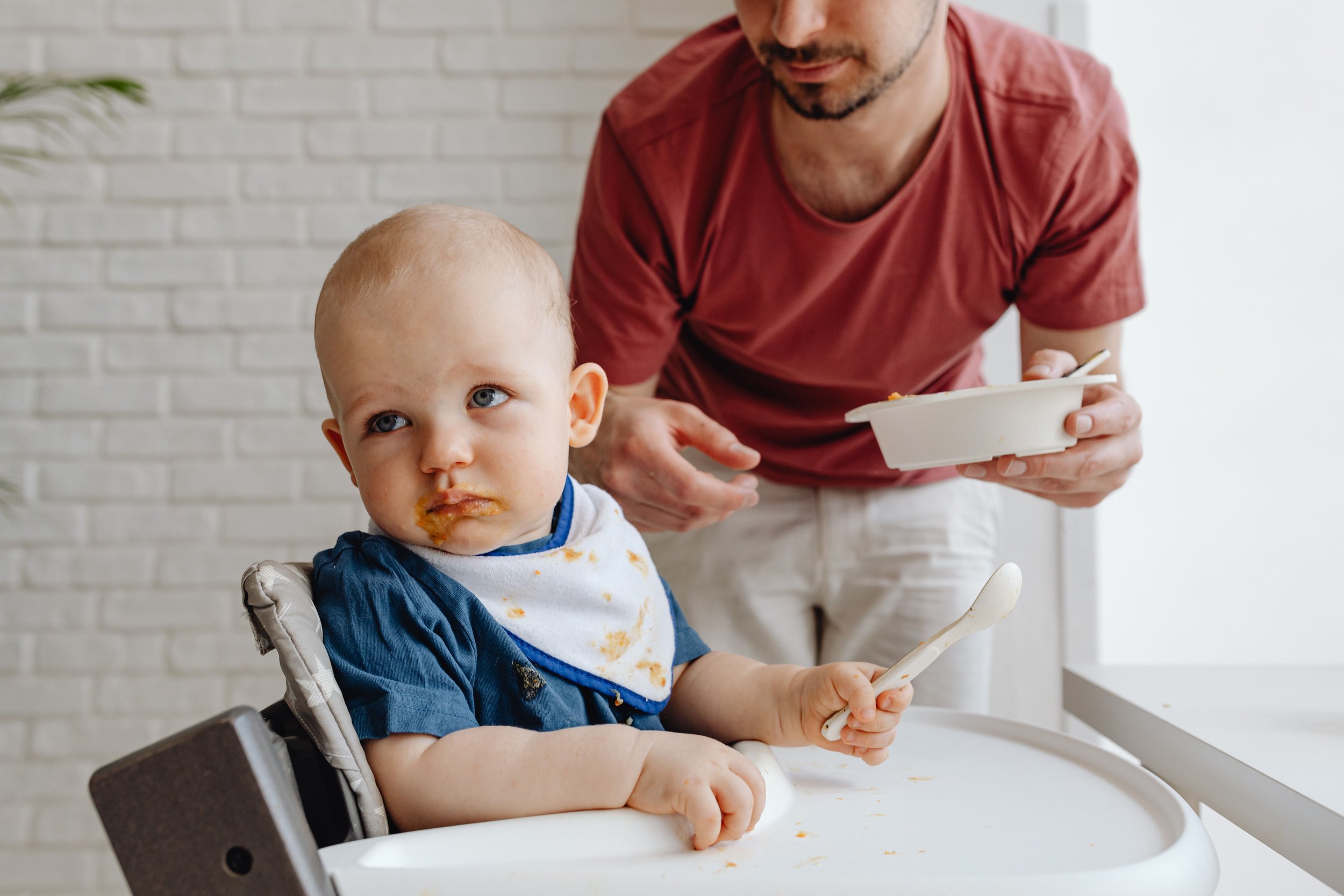 A young child with blue eyes sitting in a high chair, eating food, with some food around their mouth, wearing a bib, and holding a spoon, while an adult male with a beard and red shirt leans over nearby holding a bowl.