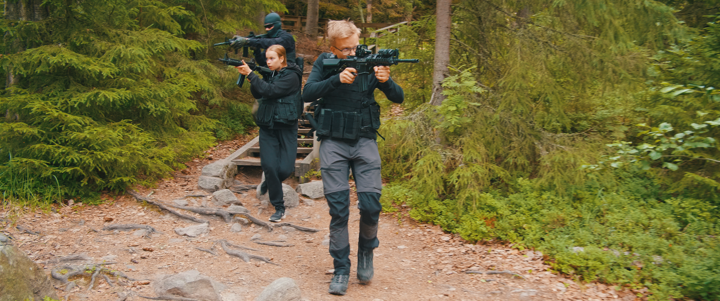 Kolme ihmistä lähtee metsäpolkua pitkin puolijohteisiin varusteisiin pukeutuneina, kantavat aseita ja liikkuvat varovasti metsän keskellä, Charles Smith - Koston Kyynel -elokuva, kuvakaappaus elokuvasta