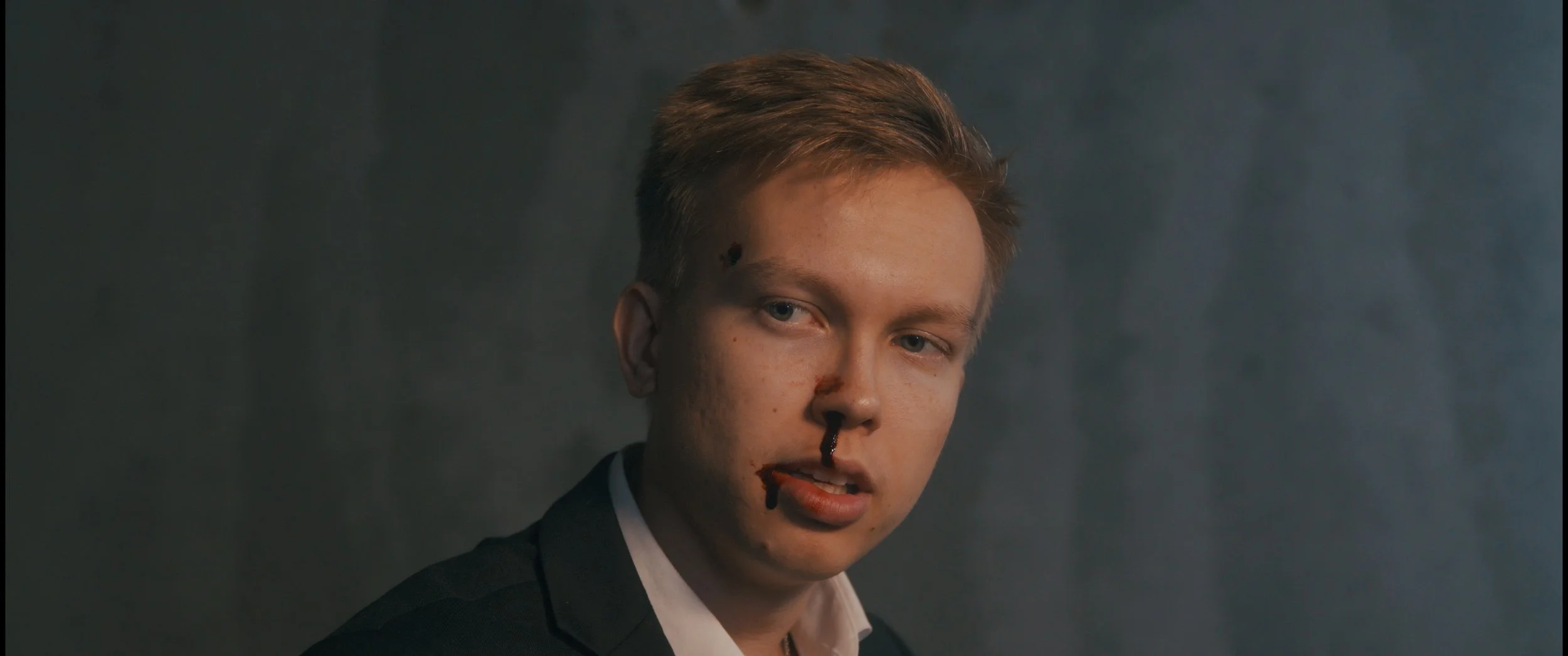 Nuori mies, jolla on verta nenässä ja huulessa, katsoo suoraan eteenpäin. Tausta harmaa. Charles Smith - Koston Kyynel -elokuva, kuvakaappaus elokuvasta, Media Aaro, Aaro Riponiemi